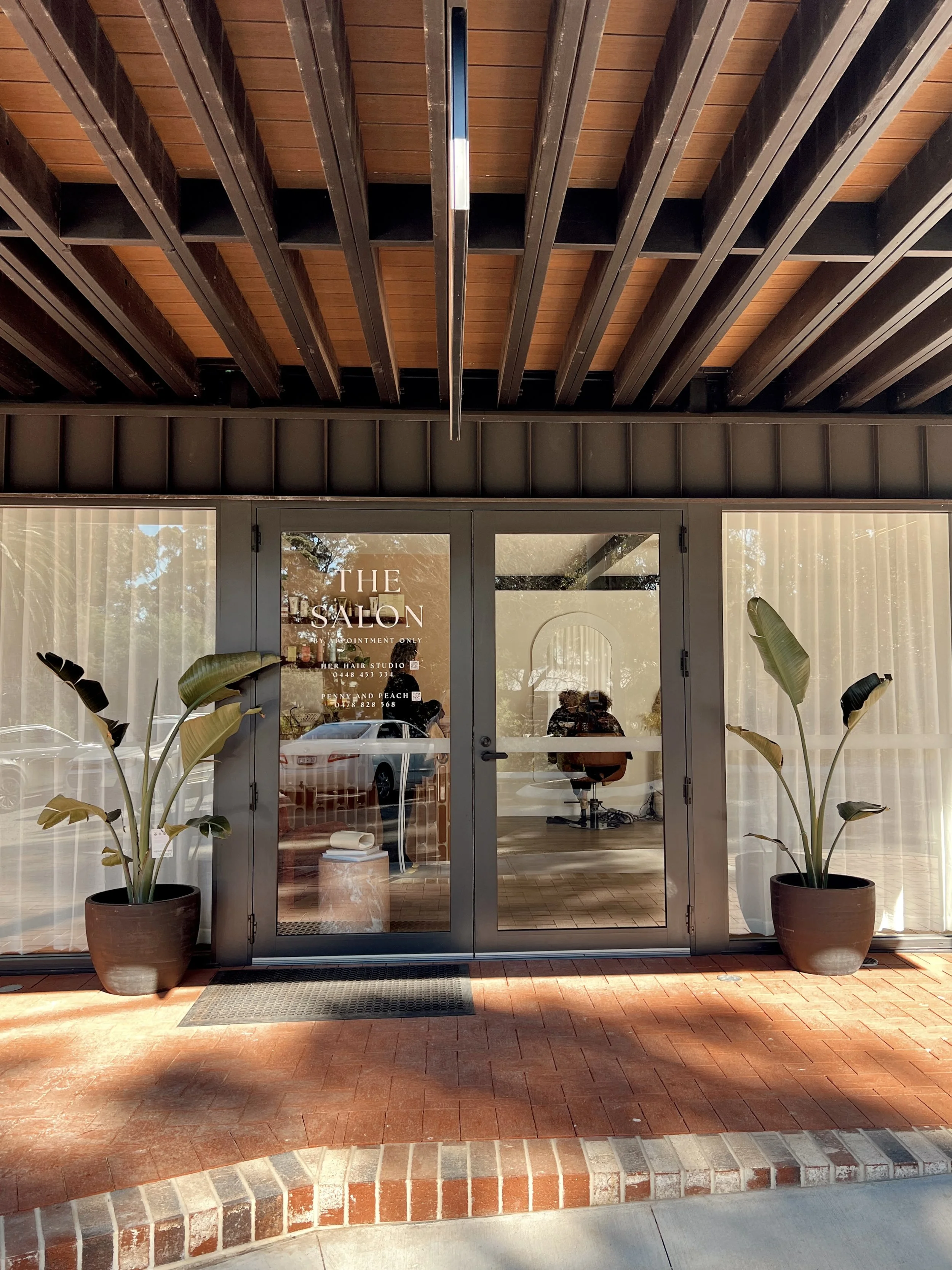 The entrance to a hair salon named 'The Salon' with glass double doors, two large potted plants on either side, and a brick sidewalk in front. Through the glass, chairs, mirrors, and some interior decor are visible.