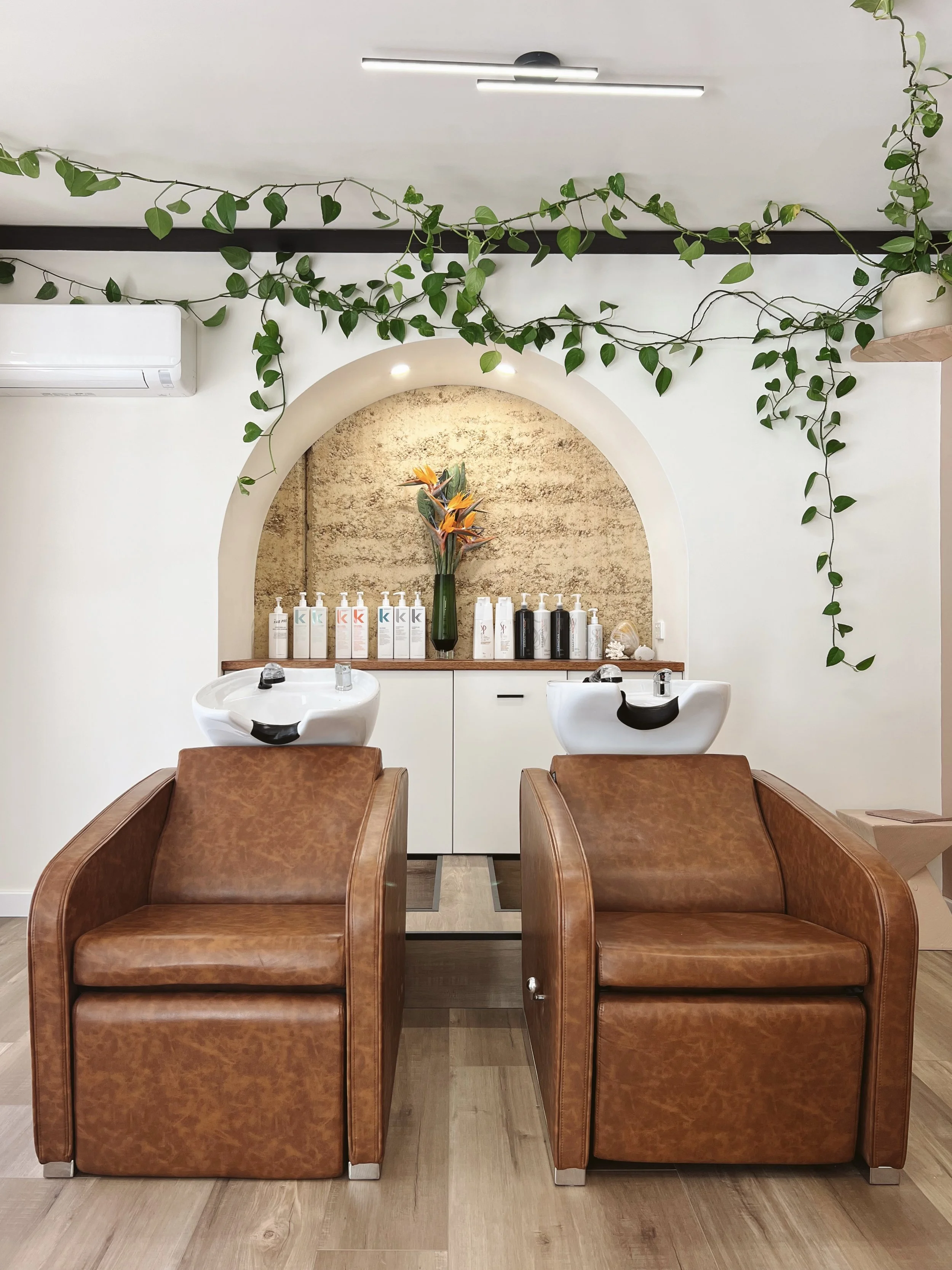 Two brown leather chairs in a salon with white sinks and various hair products on a shelf behind them, with a decorative vase of orange flowers and green plants on the wall.