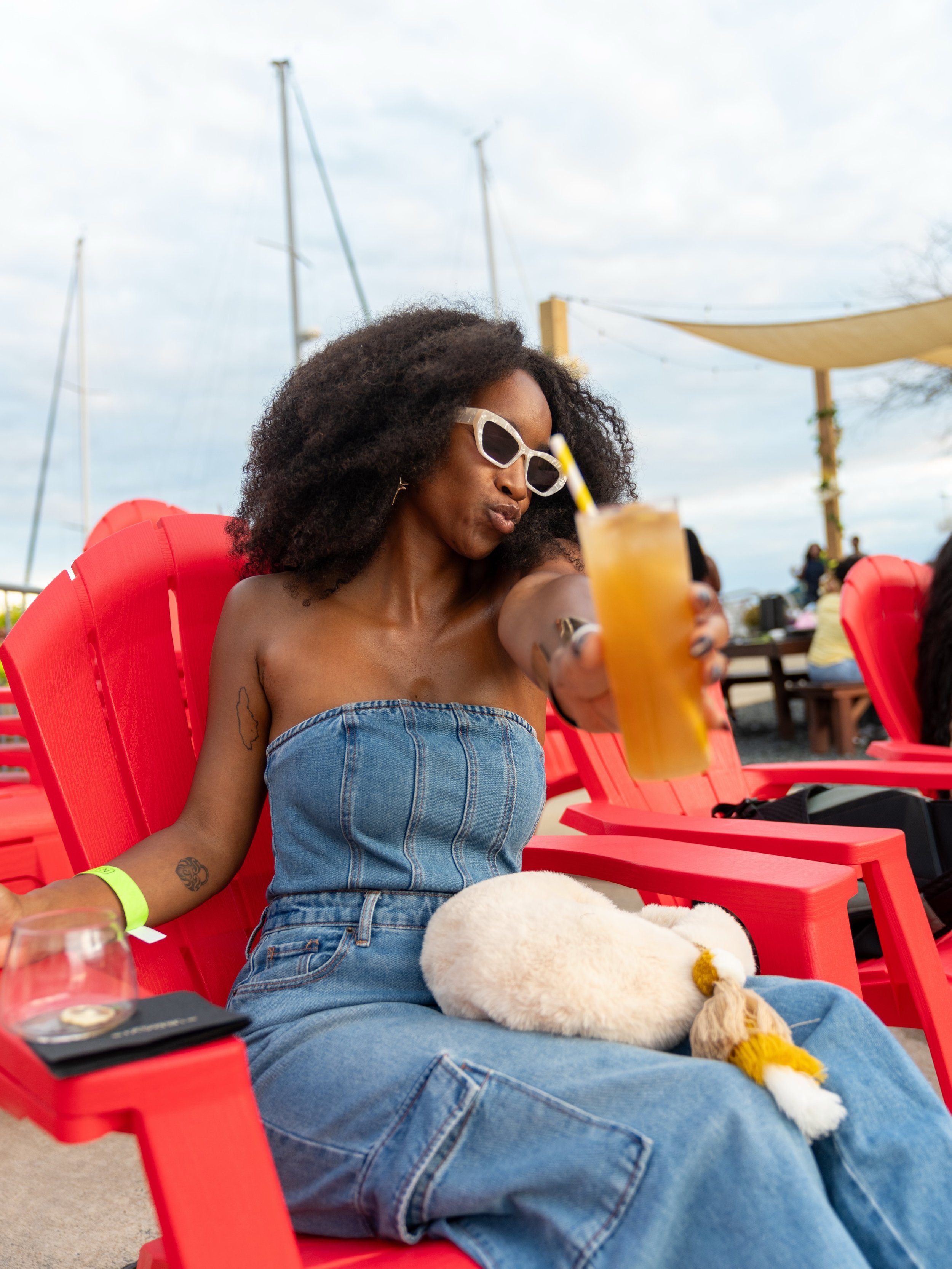 A woman with curly hair wearing sunglasses and a denim strapless top sitting on a red chair, holding a yellow drink with a straw. She has tattoos on her arm and a yellow bracelet. There is a white stuffed animal on her lap. The background shows an outdoor setting with other people, umbrellas, and a cloudy sky.