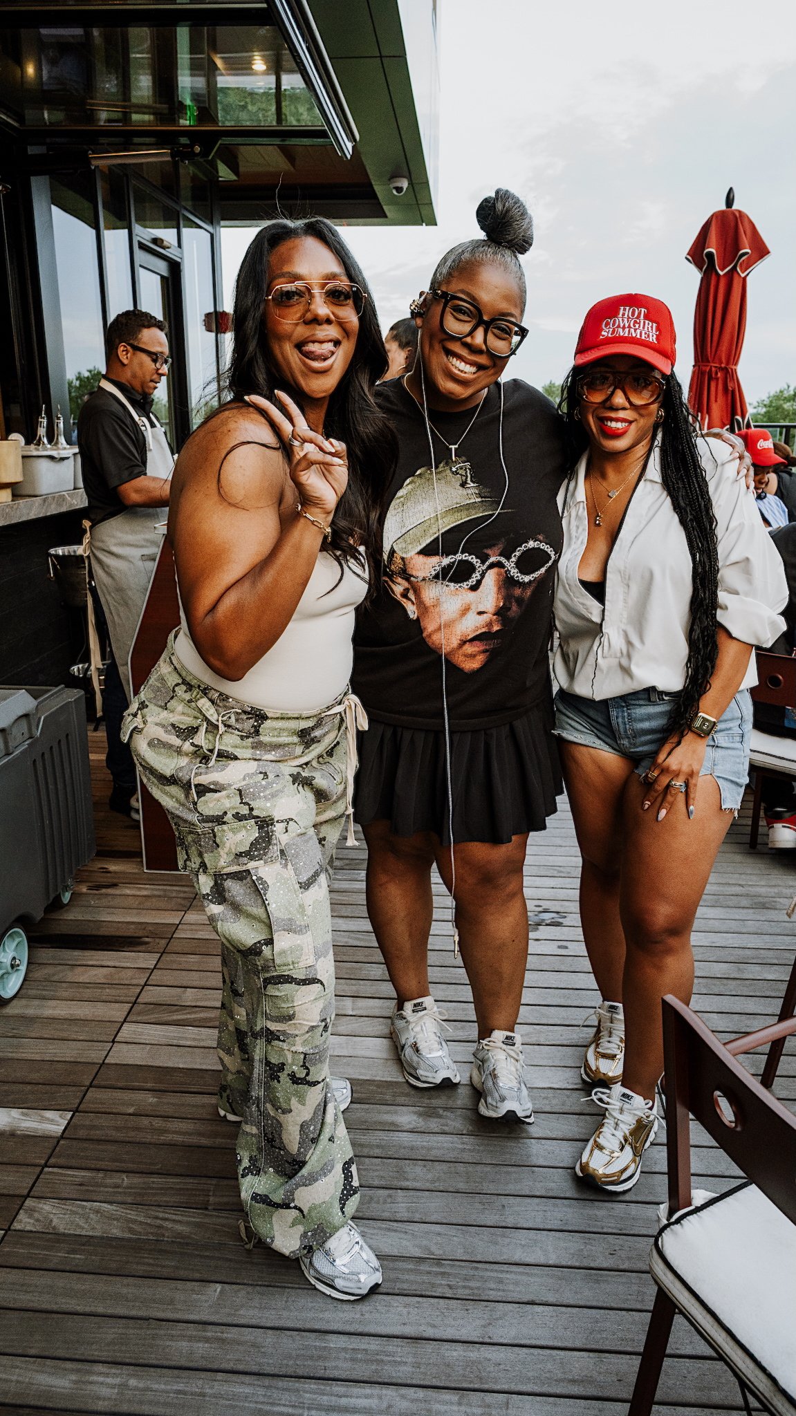Ty Alexander posing with two guests on a wooden deck at an outdoor restaurant, smiling and making playful gestures, with a man in the background preparing food.