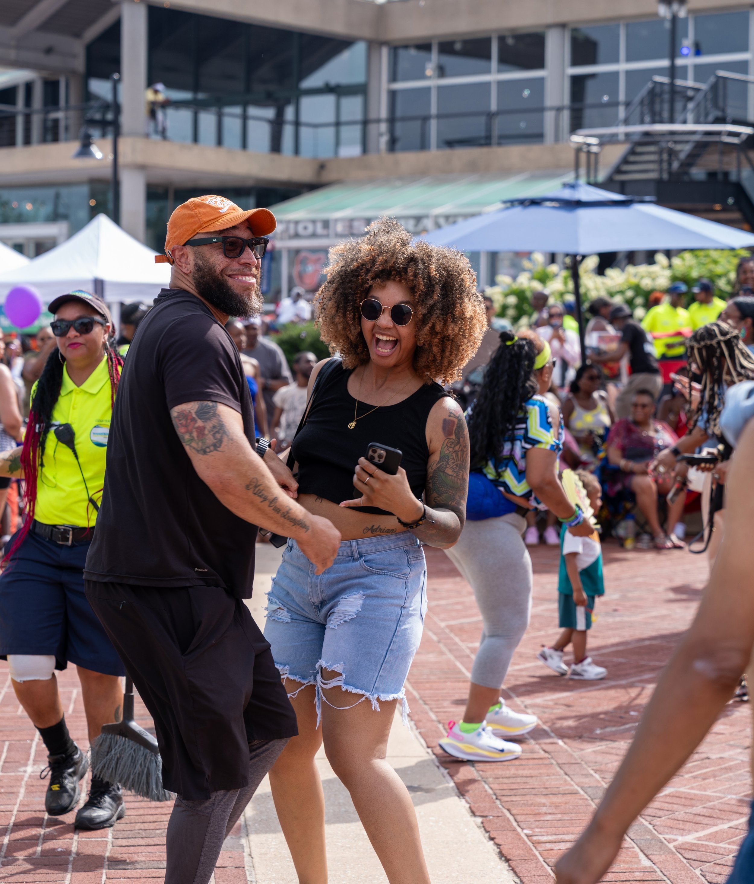 Two people smiling and interacting at an outdoor event with many people, tents, umbrellas, and a building in the background. The man wears sunglasses, an orange cap, a black t-shirt, and has tattoos on his arms. The woman has curly hair, sunglasses, a black top, and distressed denim shorts. They appear happy and engaged.