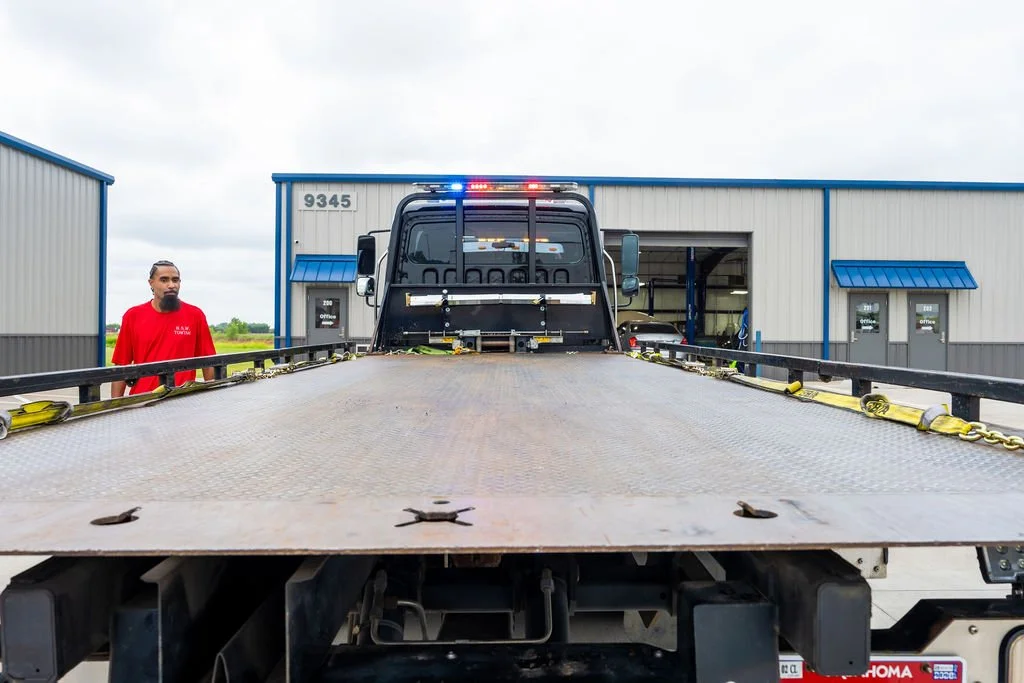 A flatbed tow truck with a black cab and emergency lights brightening the scene, parked outside a large gray industrial building with blue accents. A man in a red shirt stands nearby, and the building has the number 9345 on it.