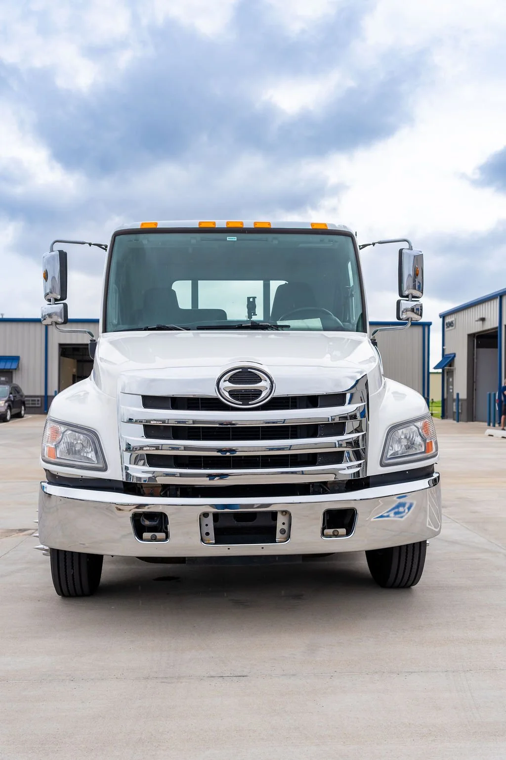 Front view of a white commercial truck parked on a concrete lot under cloudy sky, with industrial buildings in the background.