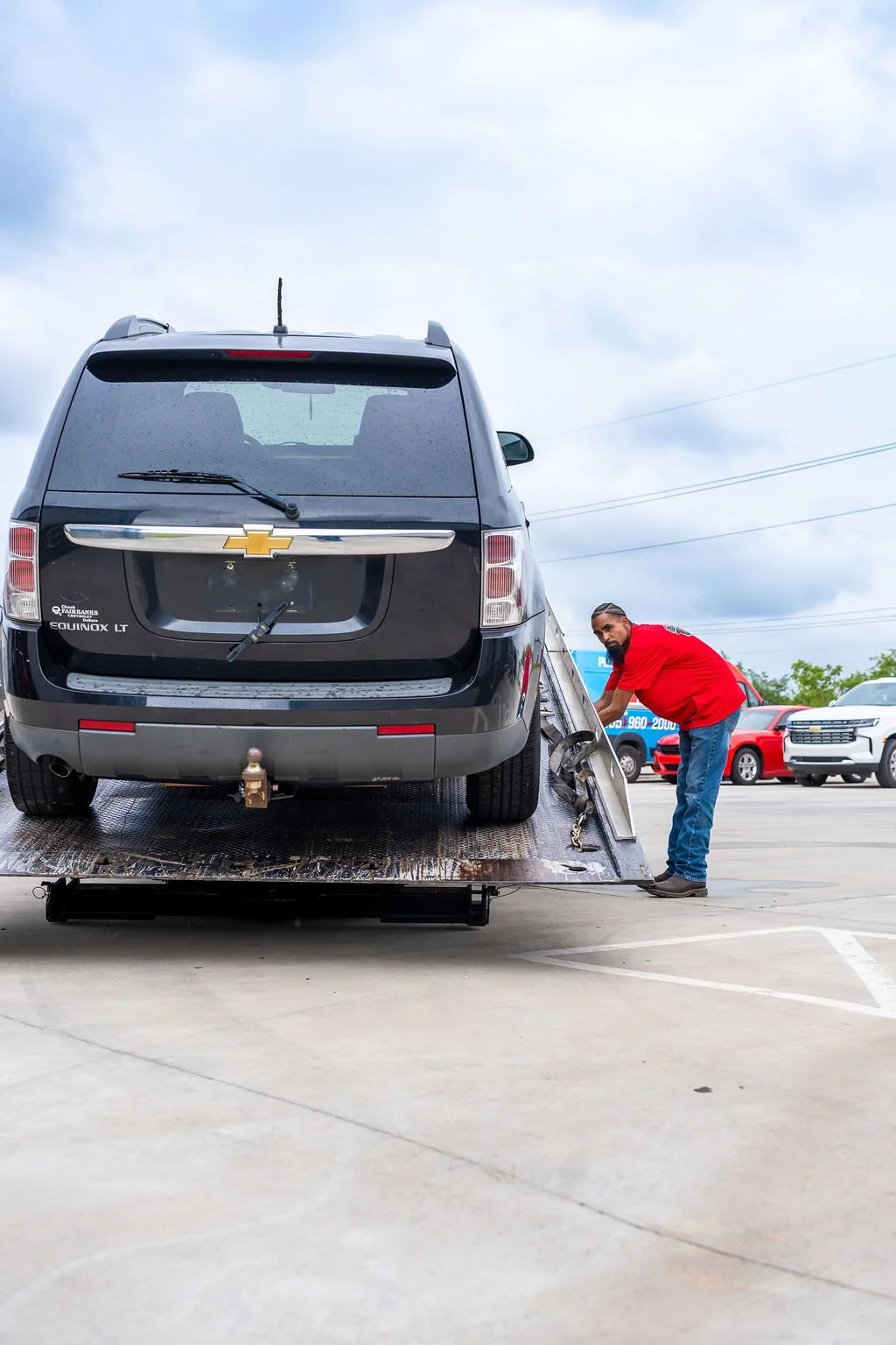 A man in a red shirt loading a black Chevrolet SUV onto a flatbed truck at a parking lot on a cloudy day.