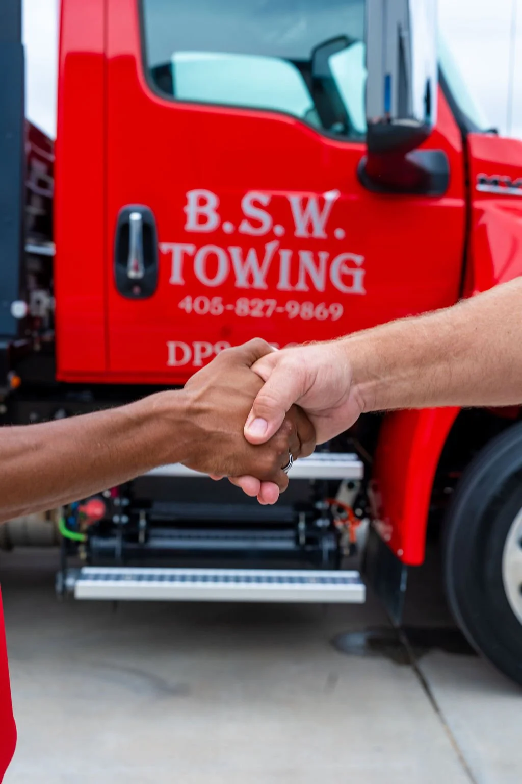 Two people shaking hands in front of a red tow truck with the text 'B.S.W. Towing' and a phone number on it.