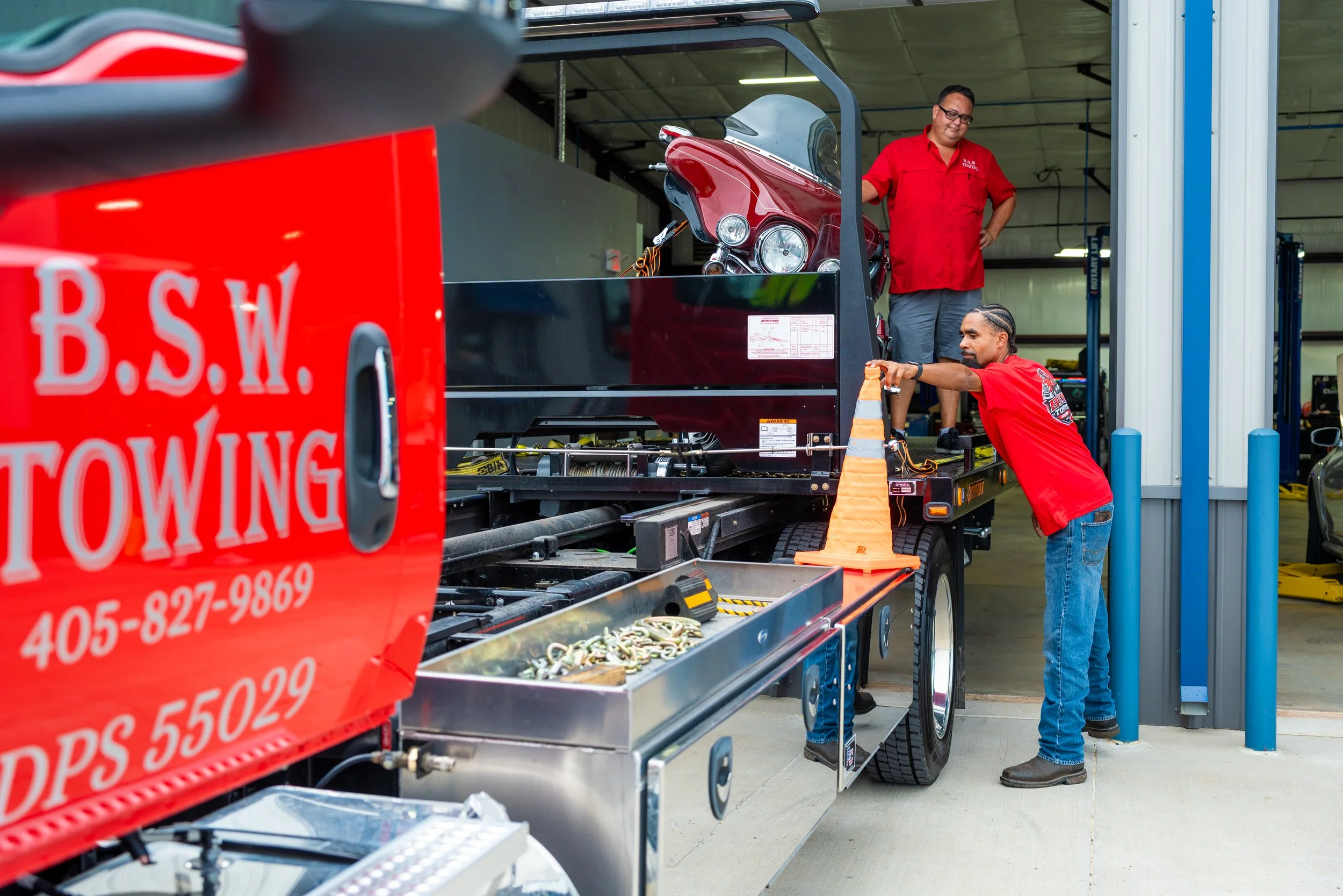 Two workers in red shirts are loading a red motorcycle into a transportation trailer. The trailer is marked with red and white lettering, and the scene takes place inside a warehouse or garage with blue safety barriers.