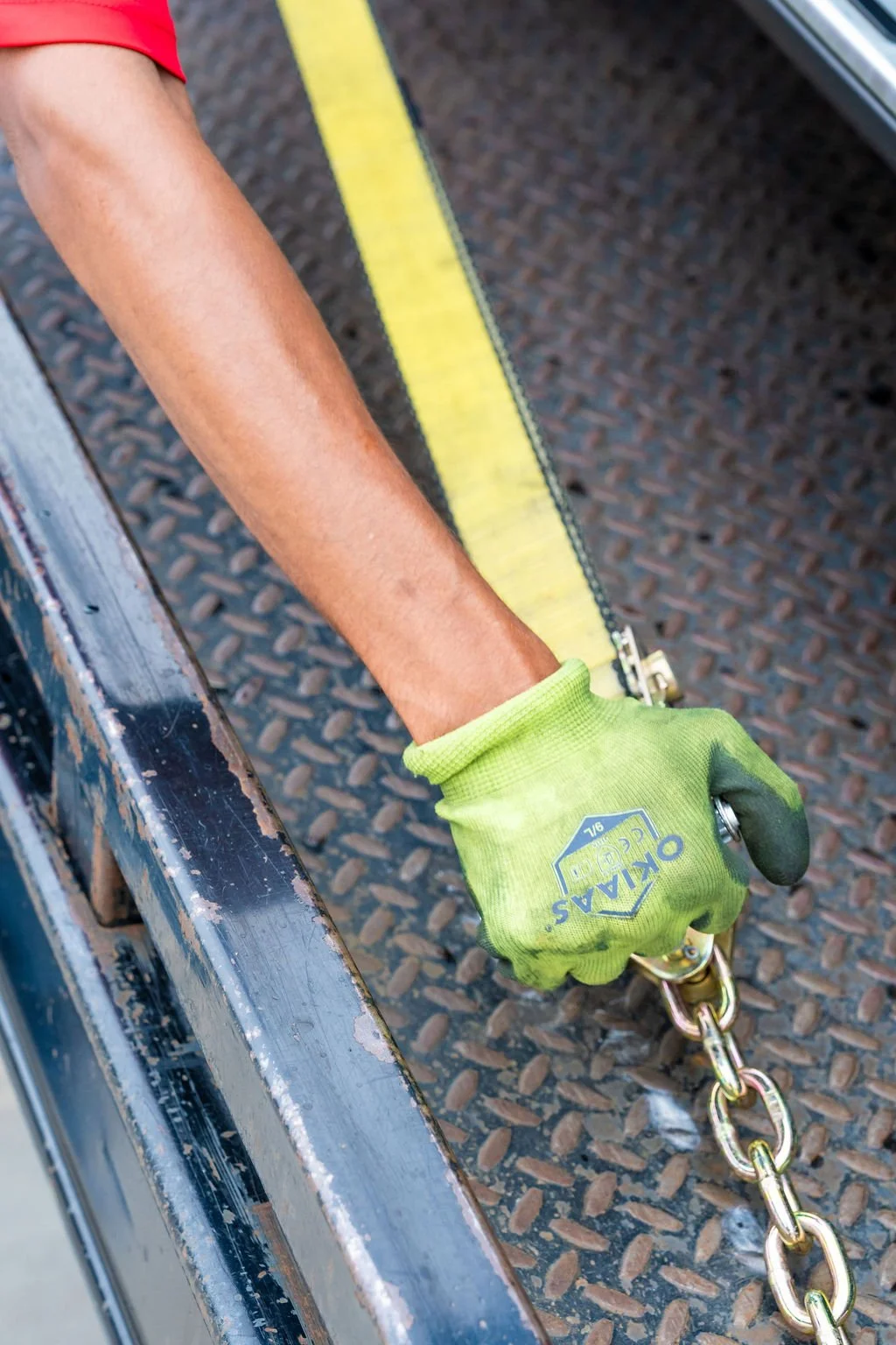 Worker wearing a green glove holds a chain attached to a yellow measuring tape, with a rusty metal surface and textured black flooring in the background.