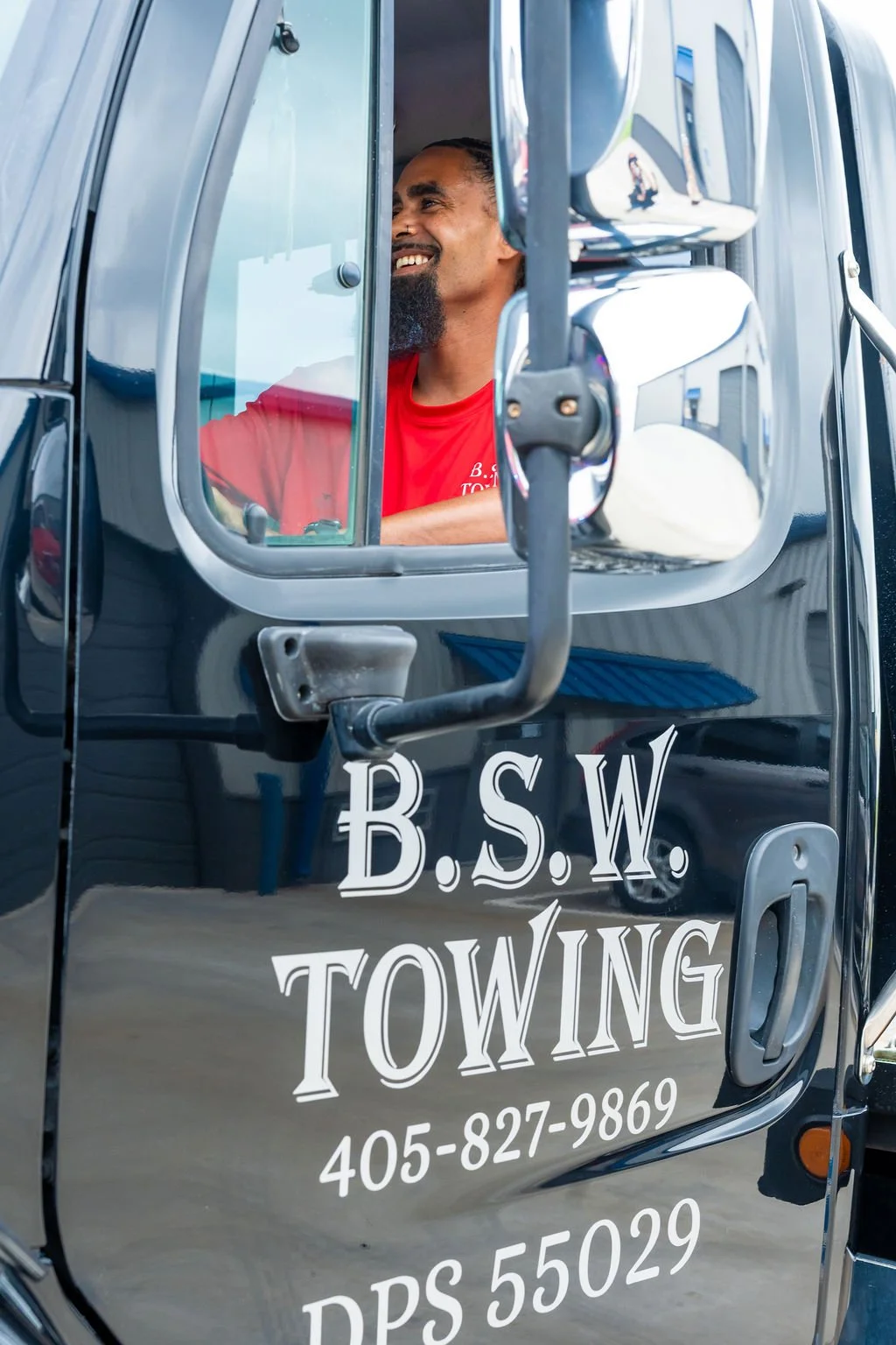A man smiling inside a black tow truck with the text "B.S.W. Towing," a phone number, and a DOT number written on the door.