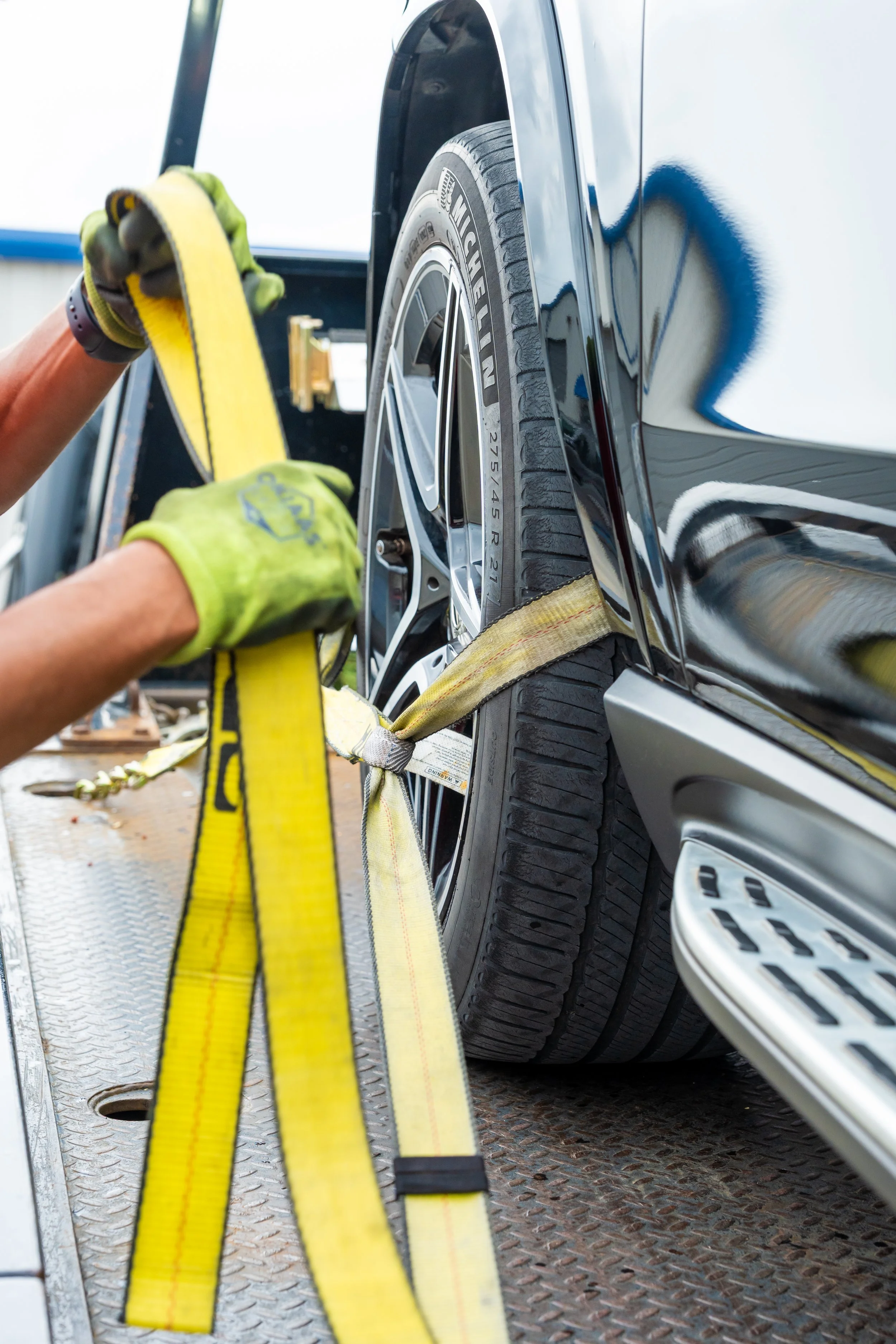 A person in gloves is securing a black car to a flatbed tow truck using a yellow strap.