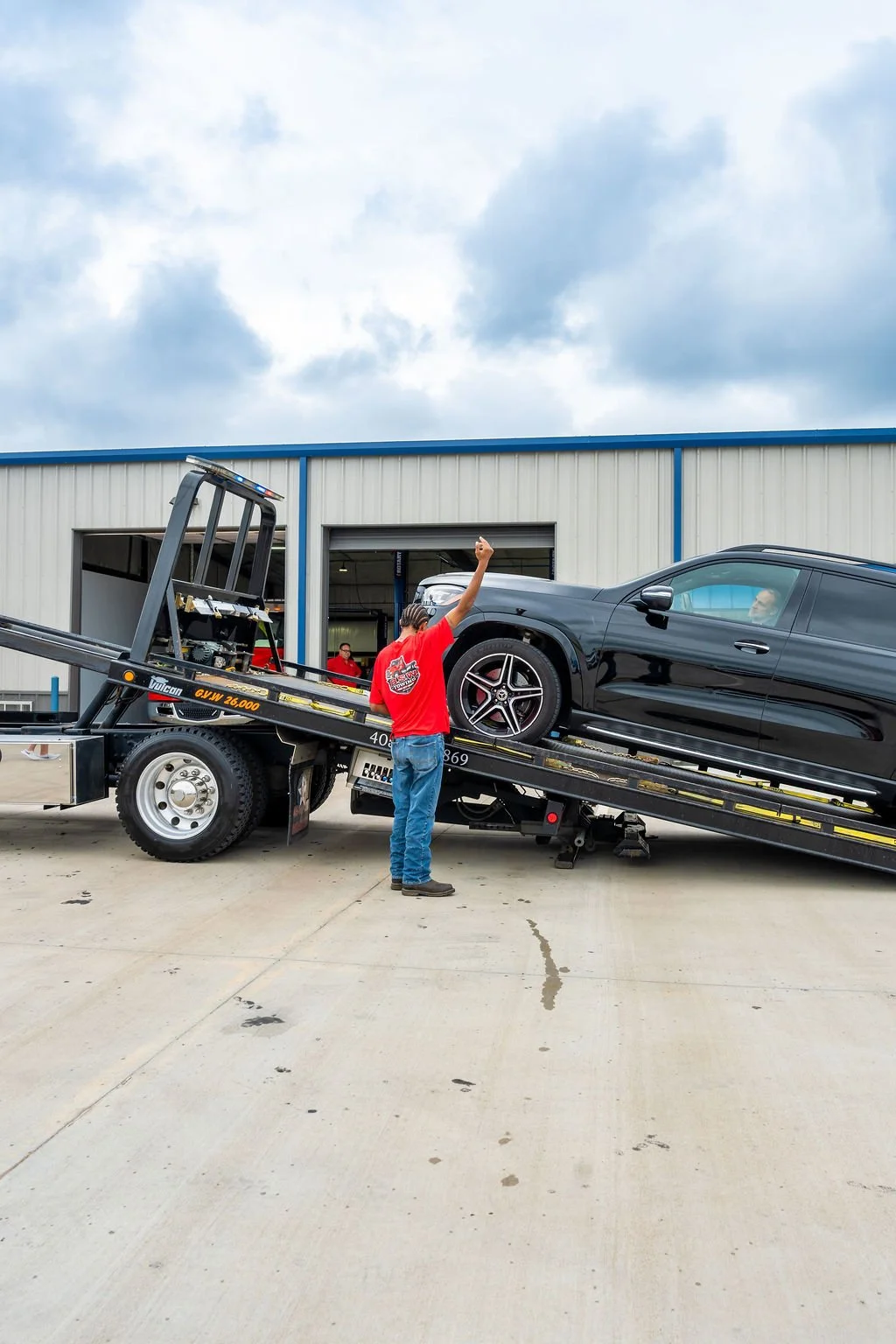 A person with a red shirt and jeans raising their fist while standing next to a black car on a tow truck outside a warehouse.