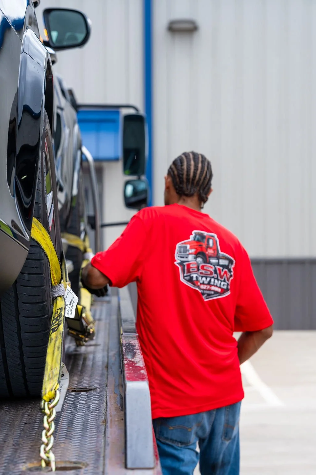 A man with cornrow braids wearing a red T-shirt with a truck logo, standing next to a black vehicle on a flatbed tow truck.