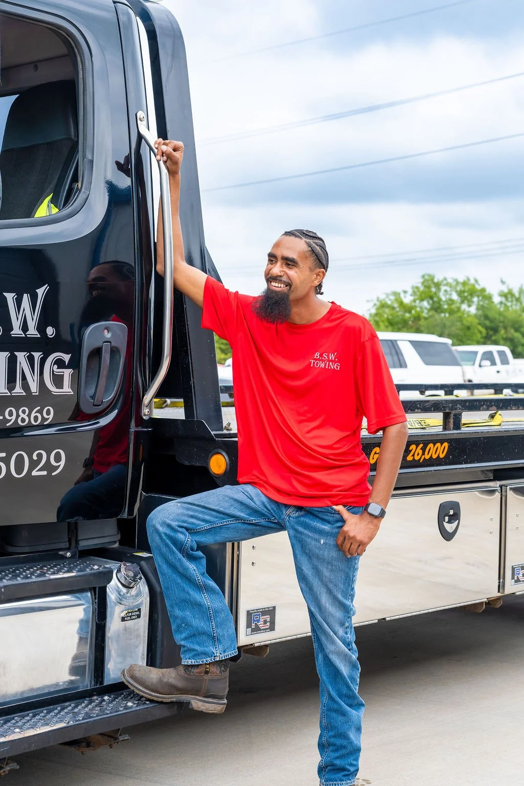 A man with a beard, wearing a red T-shirt and jeans, is smiling and leaning against a black tow truck, with one foot on the step and one hand on the door handle. The sky is cloudy, and there are other vehicles in the background.