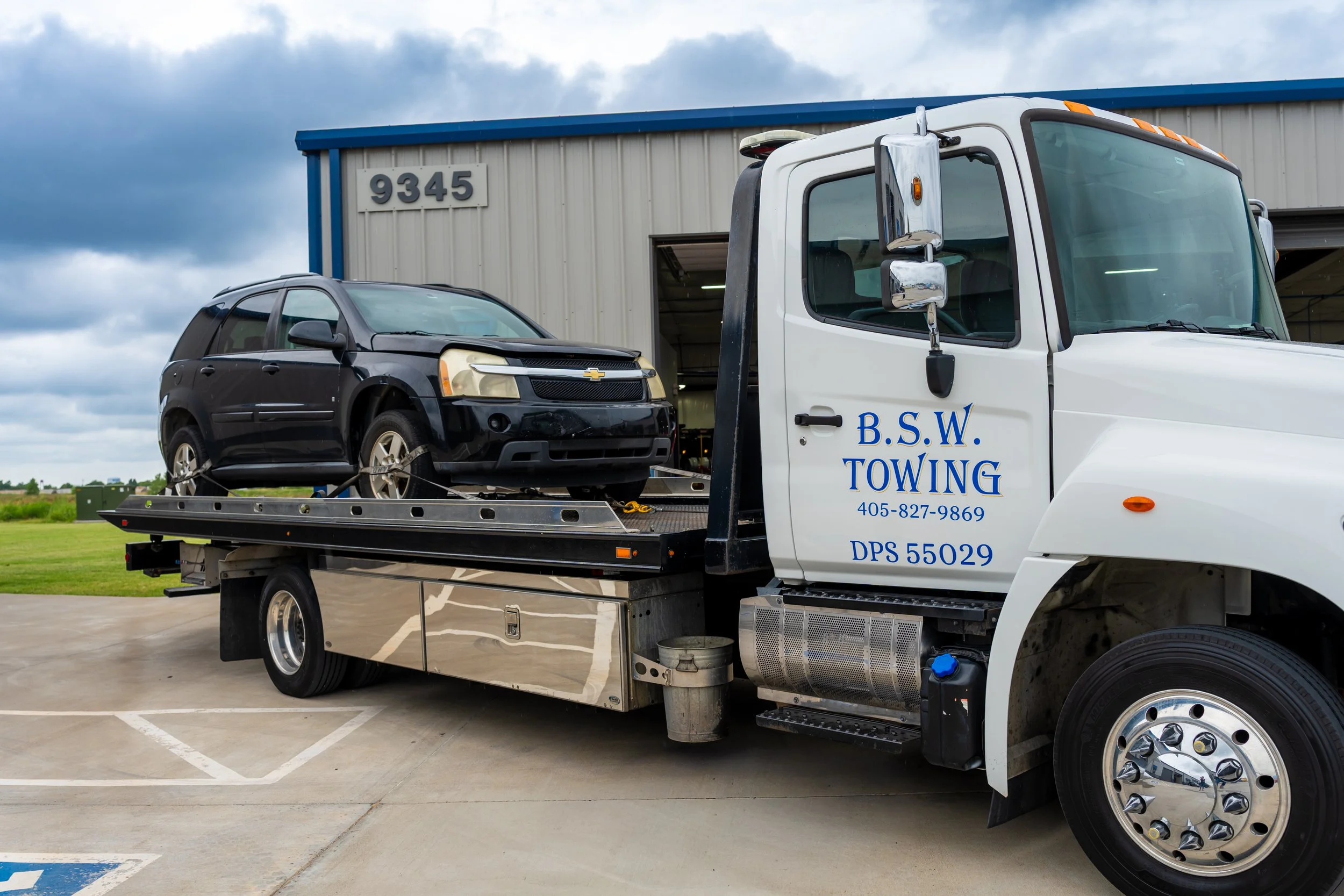 A tow truck carrying a black Chevrolet SUV in a parking lot outside a warehouse.
