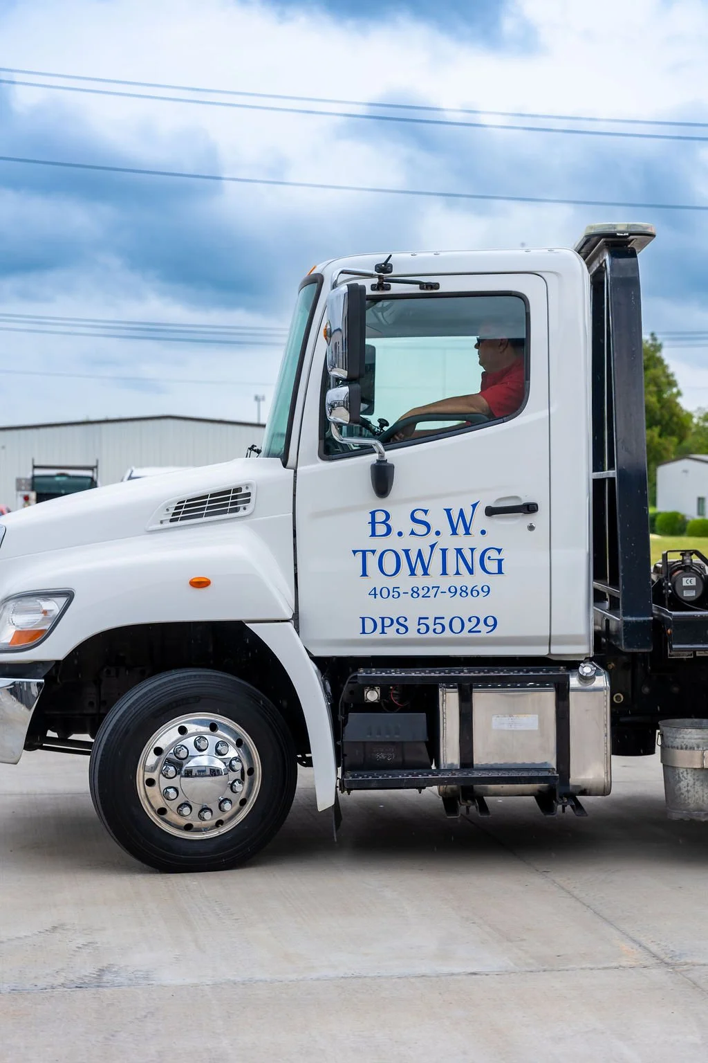 White tow truck with blue lettering that reads B.S.W. Towing, and a phone number, parked on a concrete lot under a partly cloudy sky, with a man in a red shirt inside the cab.