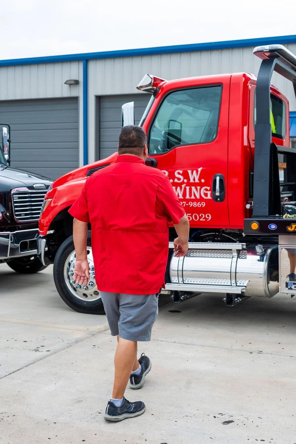 A man in a red shirt and gray shorts walking near a red tow truck marked 'S.S.W. Towing' parked outside a building.