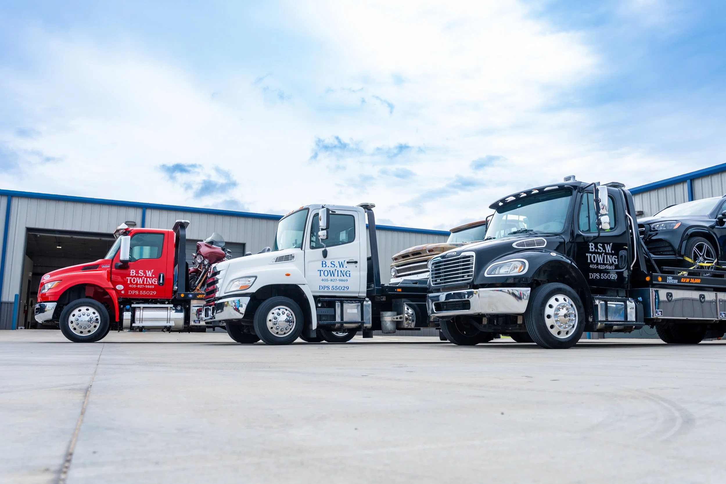 Lineup of three tow trucks parked outside a warehouse, with a partially visible black car on a flatbed, under a partly cloudy sky.