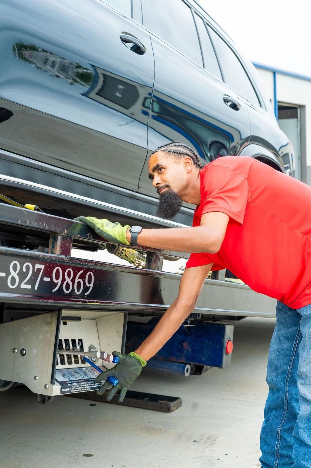 A man wearing a red shirt and green gloves is working on a flatbed truck used to transport a black car. He is checking or adjusting something on the underside of the vehicle, possibly during a maintenance or repair process.