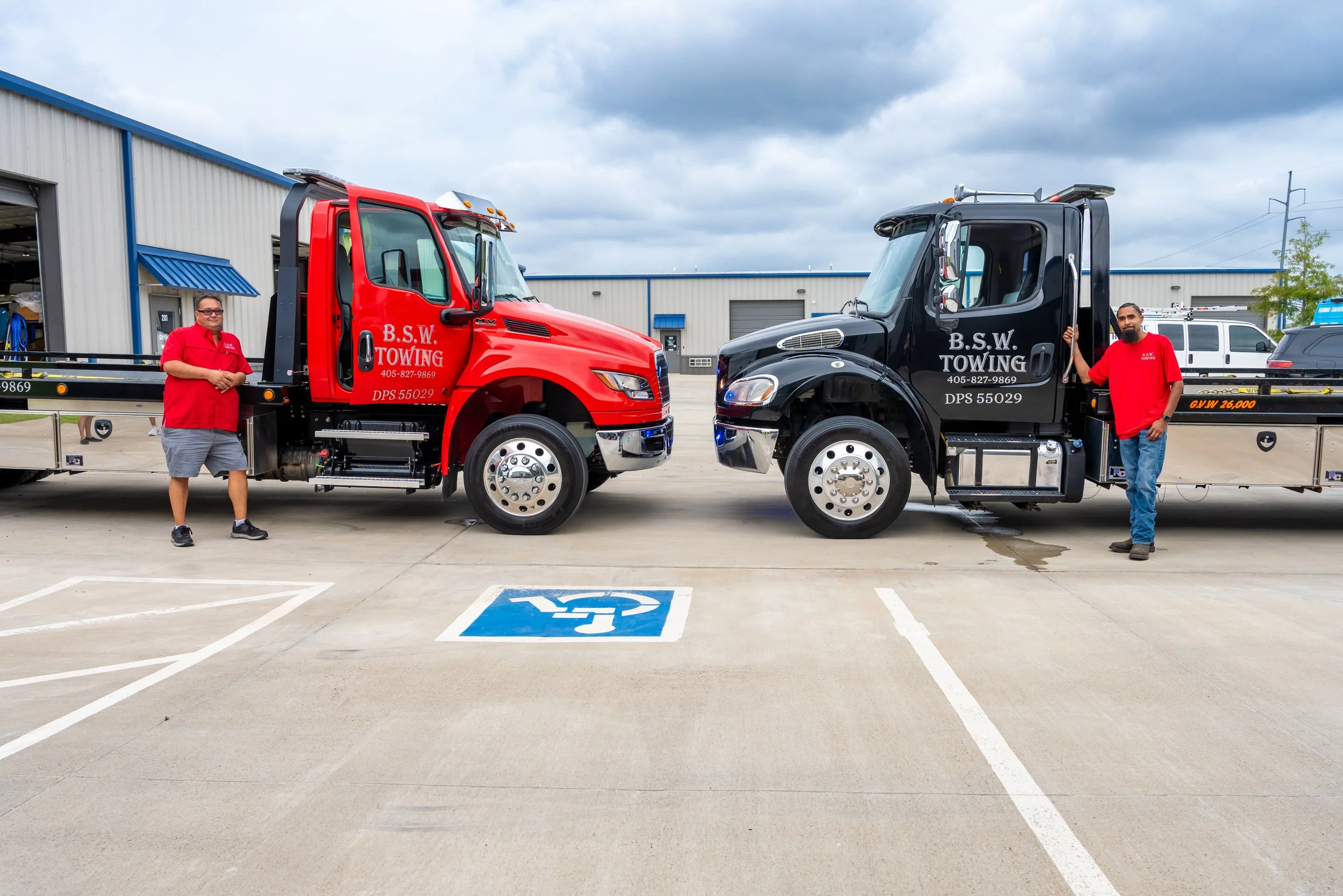 Two tow trucks, a red one and a black one, are parked side by side. Two men, one in a red shirt and the other in a red polo shirt, are standing near the trucks. The trucks have the company's name "B.S.W. Towing" written on them. The scene is outside a building with a cloudy sky overhead, and a marked handicapped parking space is visible in the foreground.