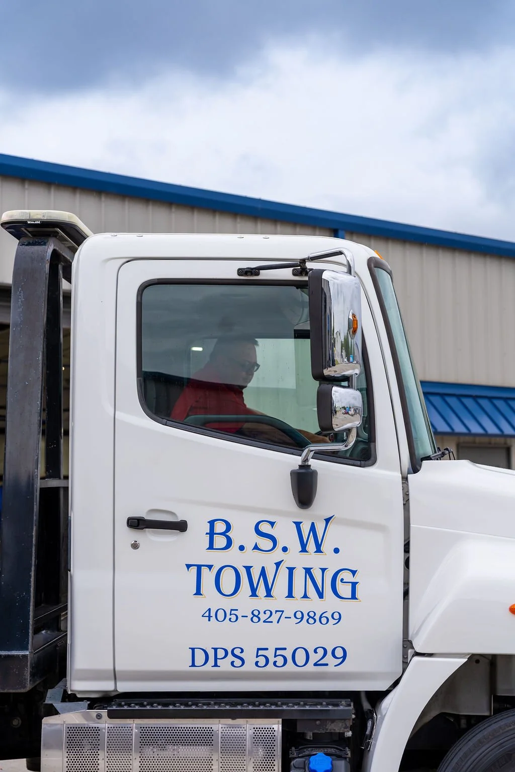 Close-up of a white tow truck with the business name B.S.W. Towing, phone number 405-827-9869, and DPS 55029 written on the side, parked outside a building with a blue roof.
