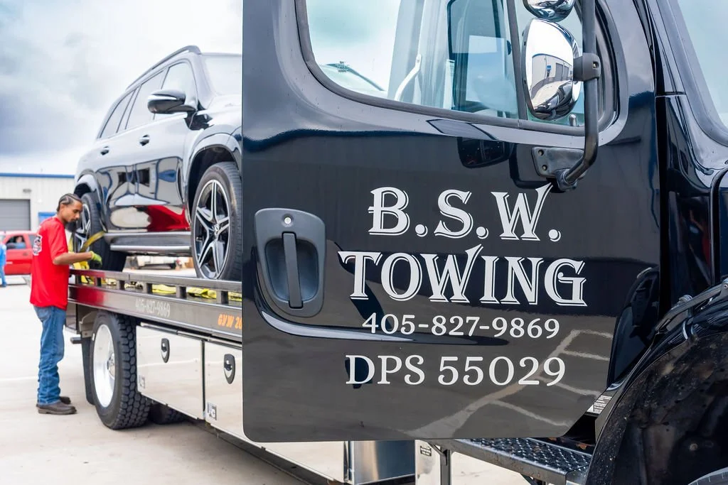 A person in a red shirt and jeans working on a black towing truck with the words "B.S.W. Towing" and a phone number on the door. The truck is towing a black SUV. There are other vehicles and a building in the background.
