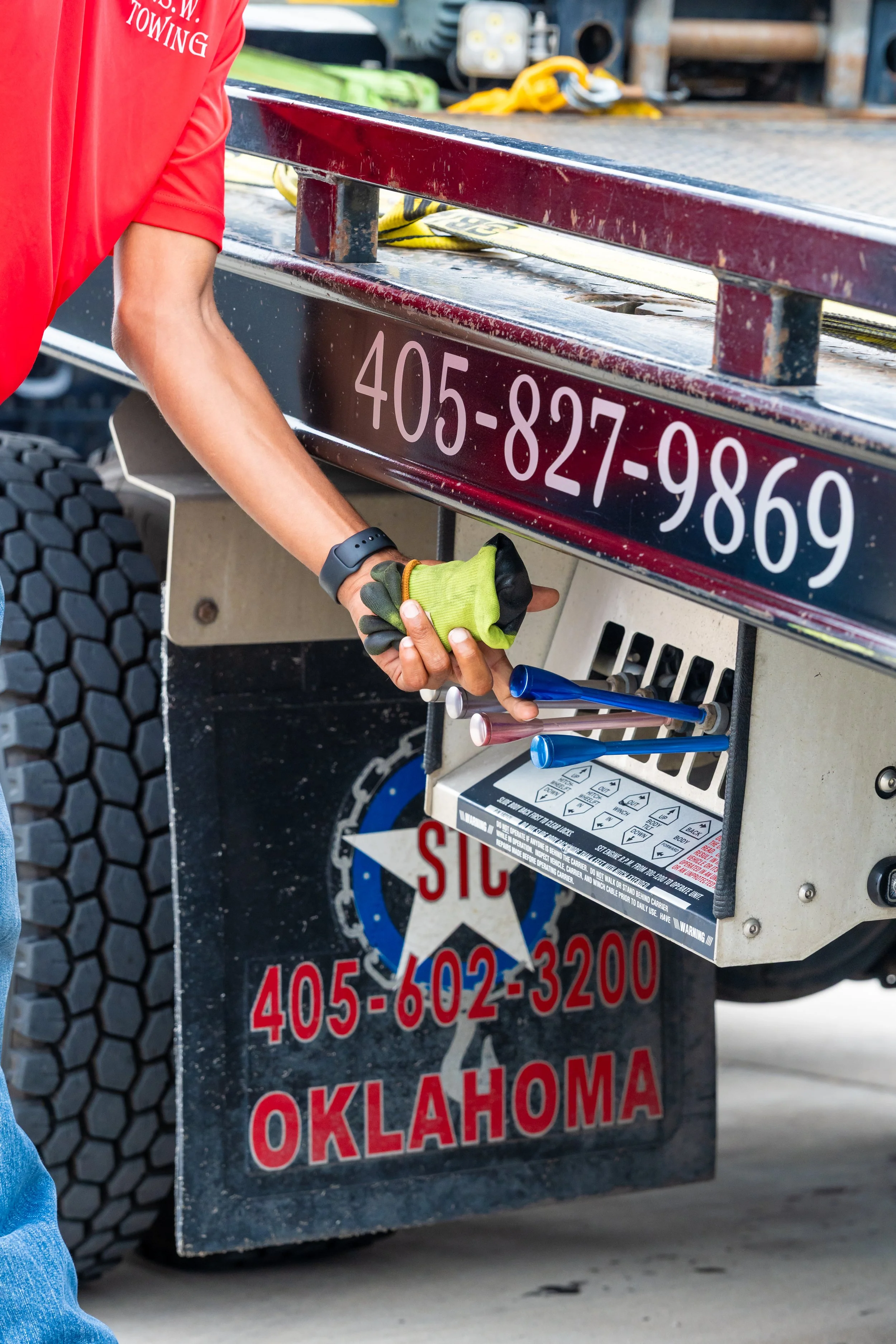 Close-up of a person securing a tow truck with a green and black glove, with a red shirt, on a concrete surface. The truck has a black, textured tire and a front bumper with a safety warning label, showing a Texas license plate with red and white text, including a phone number and the word Oklahoma.