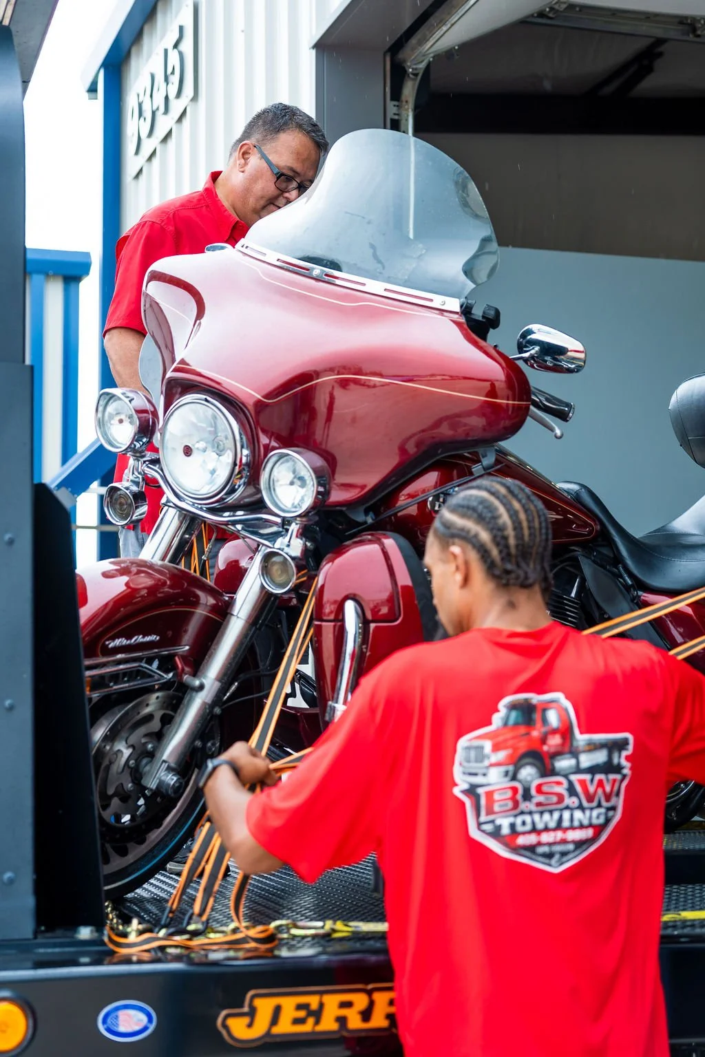 Two men in red shirts, one with cornrows, are securing a red motorcycle on a trailer with orange straps. One man is standing on the ground and the other is leaning over the motorcycle.