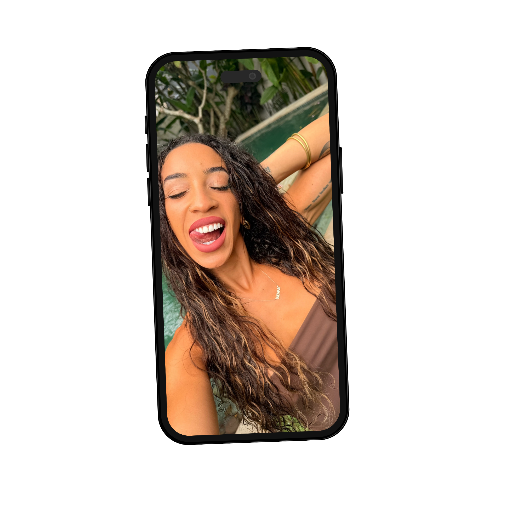 Close-up of a smiling woman with dark wavy hair, wearing hoop earrings and gold bangles, taking a selfie outdoors amidst greenery.