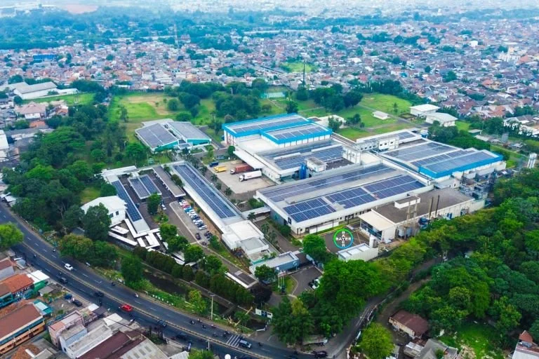 Aerial view of a large industrial complex with multiple buildings and solar panels, surrounded by green trees and a neighborhood with houses.