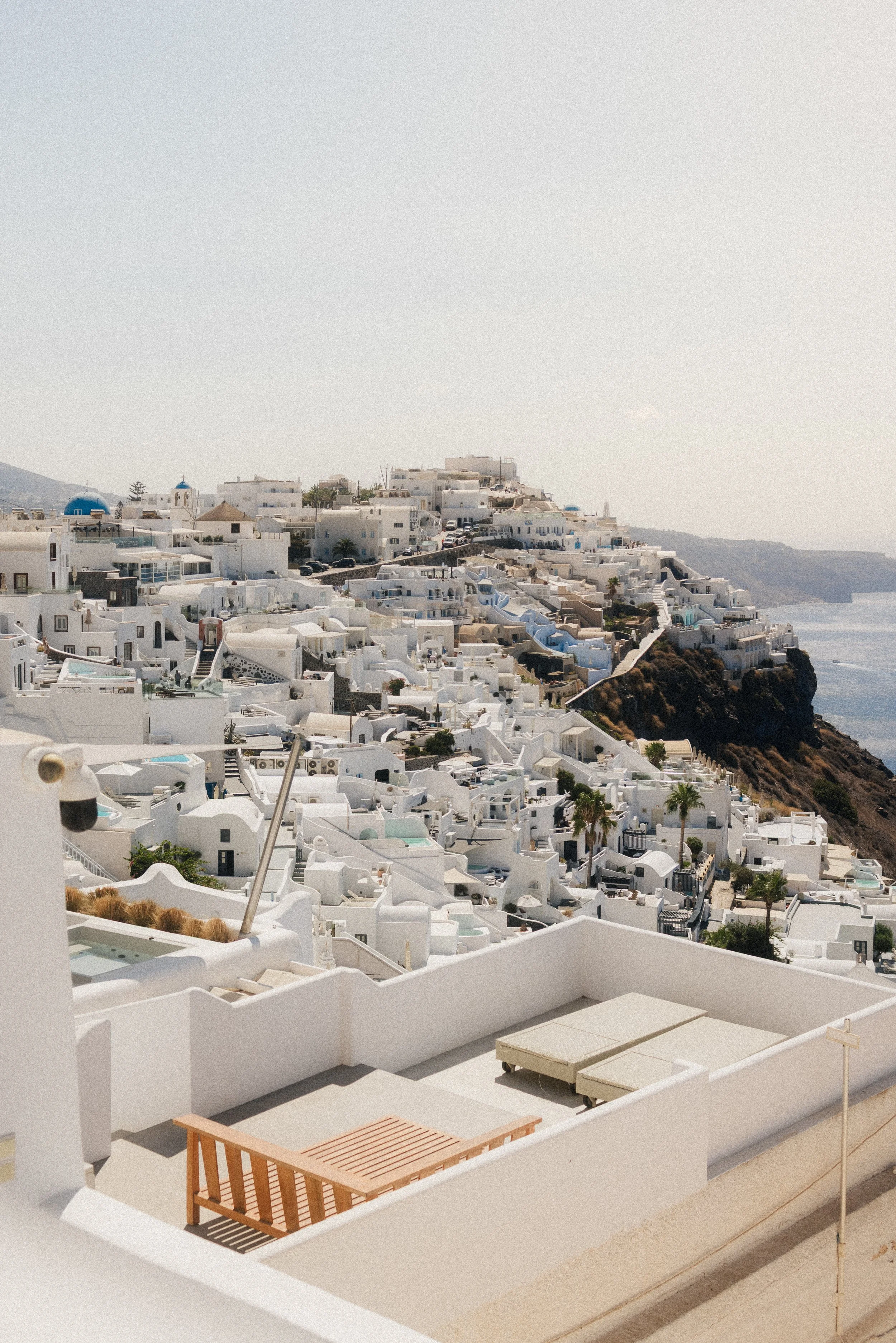 A white rooftop terrace with a wooden table and two loungers, overlooking white buildings on a hillside in Santorini, Greece, with the sea in the background.