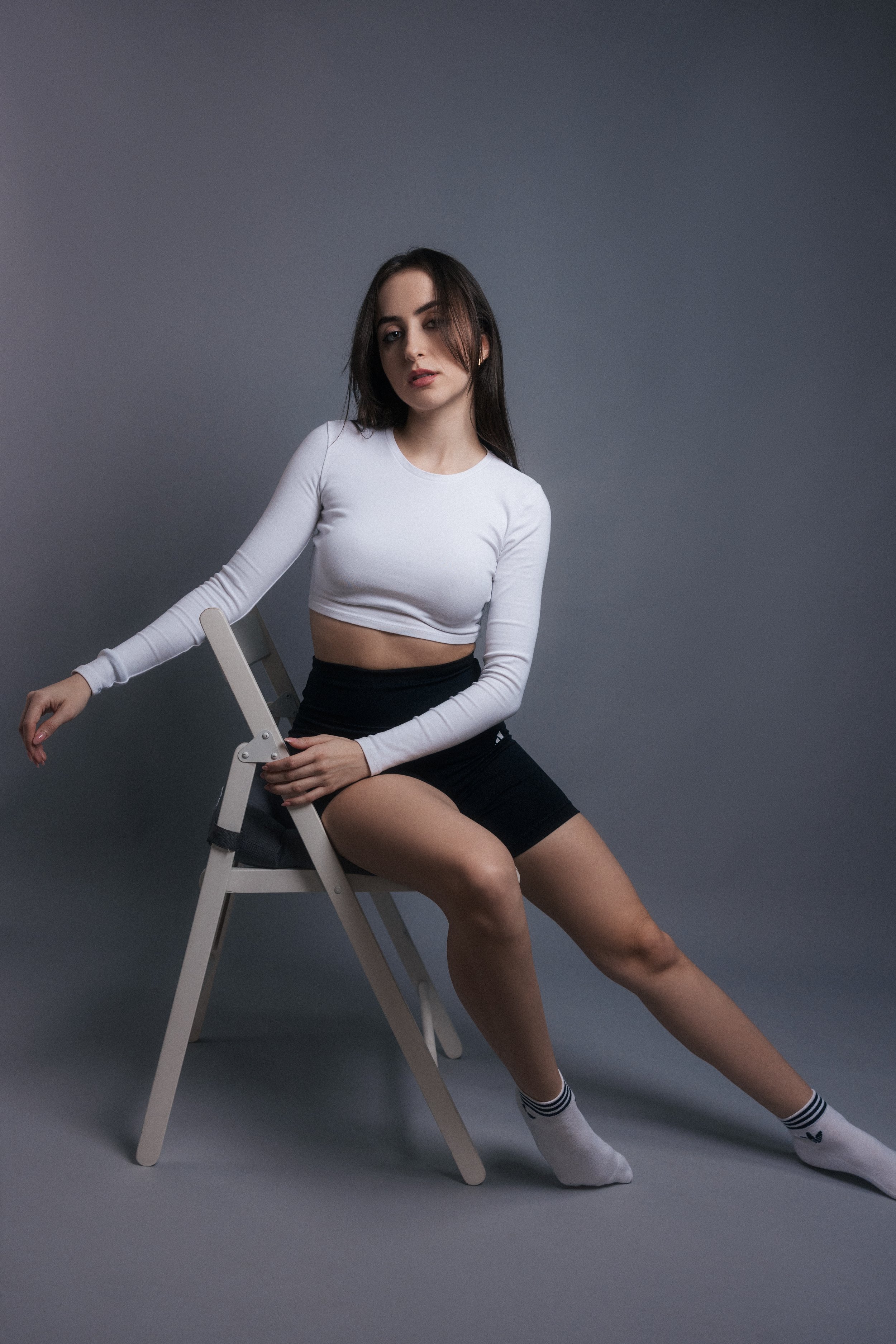 A young woman with dark hair and fair skin sitting on a tiered chair against a plain gray background, wearing a white long-sleeve crop top, black shorts, and striped socks. She is posing with one arm resting on the chair and the other on her thigh, w