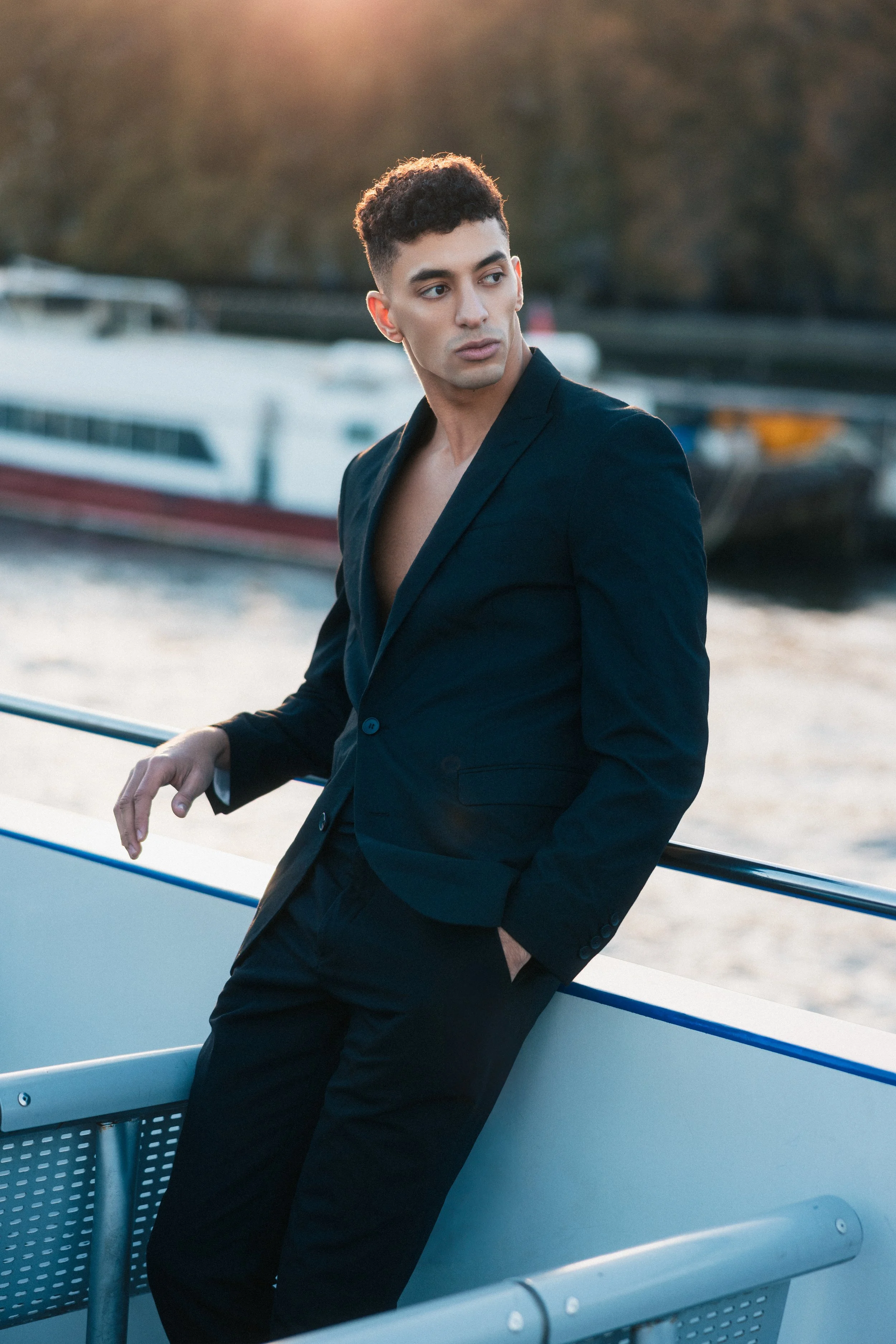 A young man with curly dark hair, wearing a black blazer and black pants, stands on a boat with a waterfront background at sunset, looking to the side with a contemplative expression.