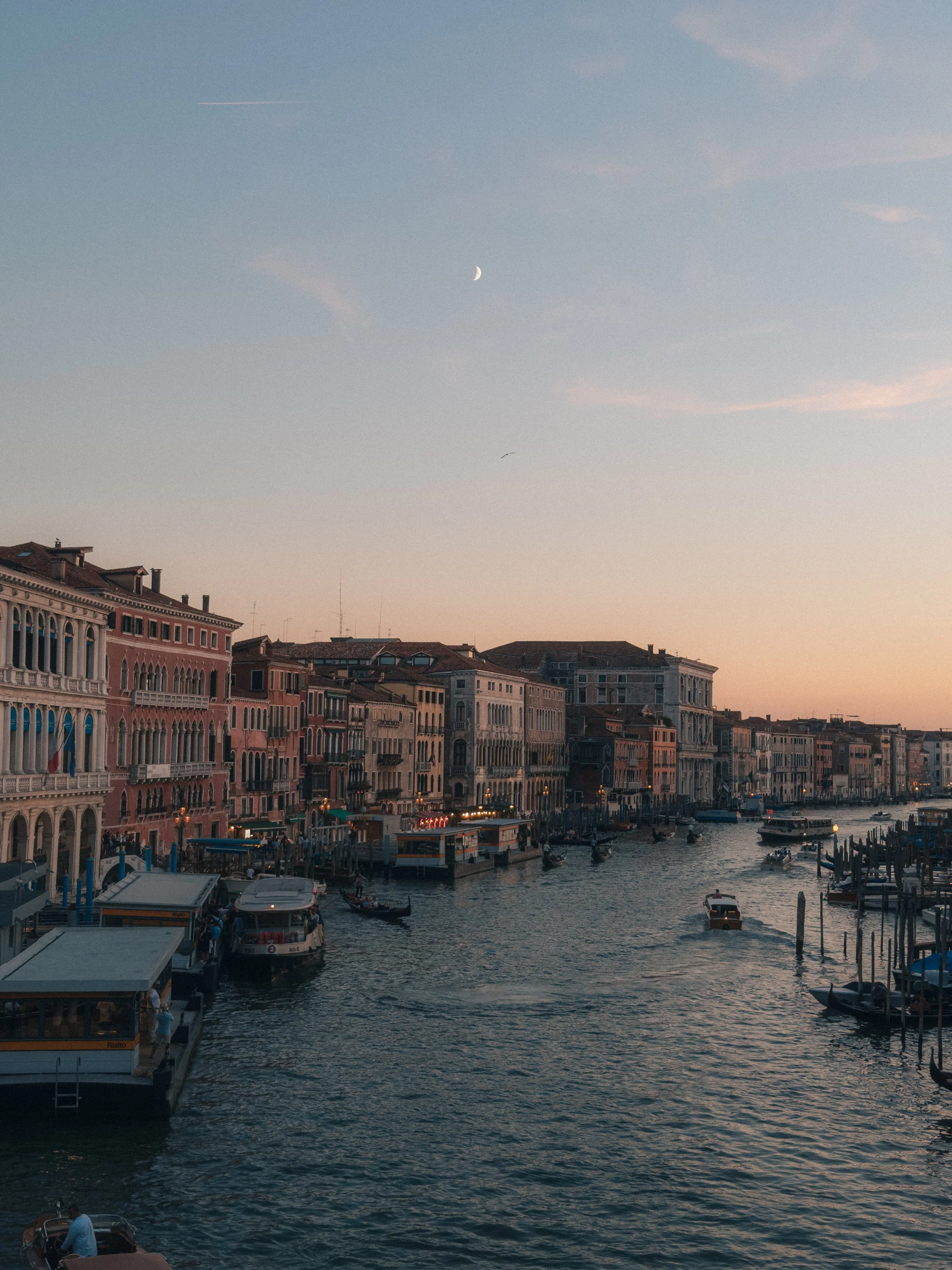 View of a canal in Venice, Italy, with historic buildings lining the water, boats docked along the sides, and a partially visible sky with a moon and clouds at dusk.