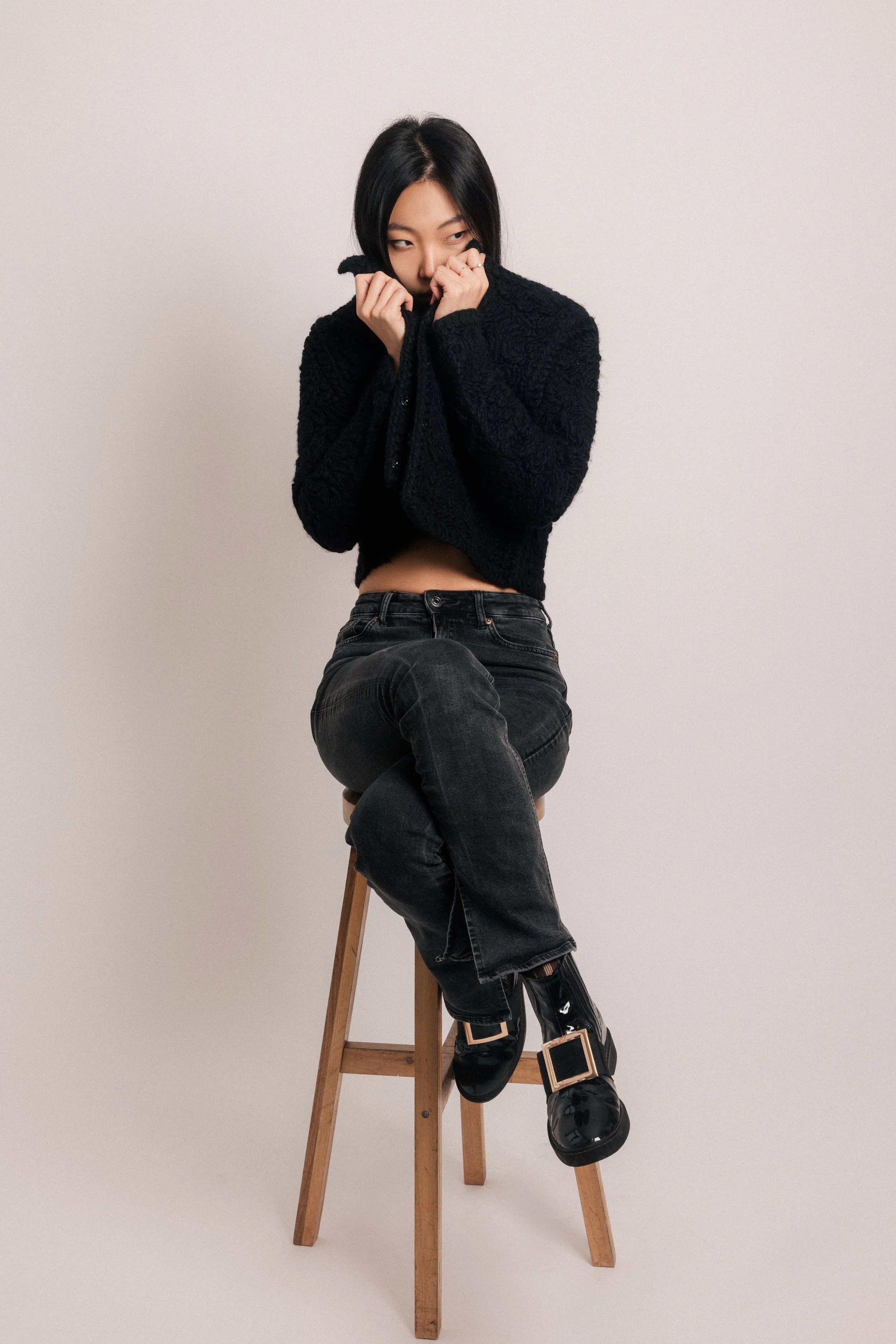 A woman with black hair wearing a black fuzzy sweater and black pants sitting on a wooden stool in front of a plain white background.