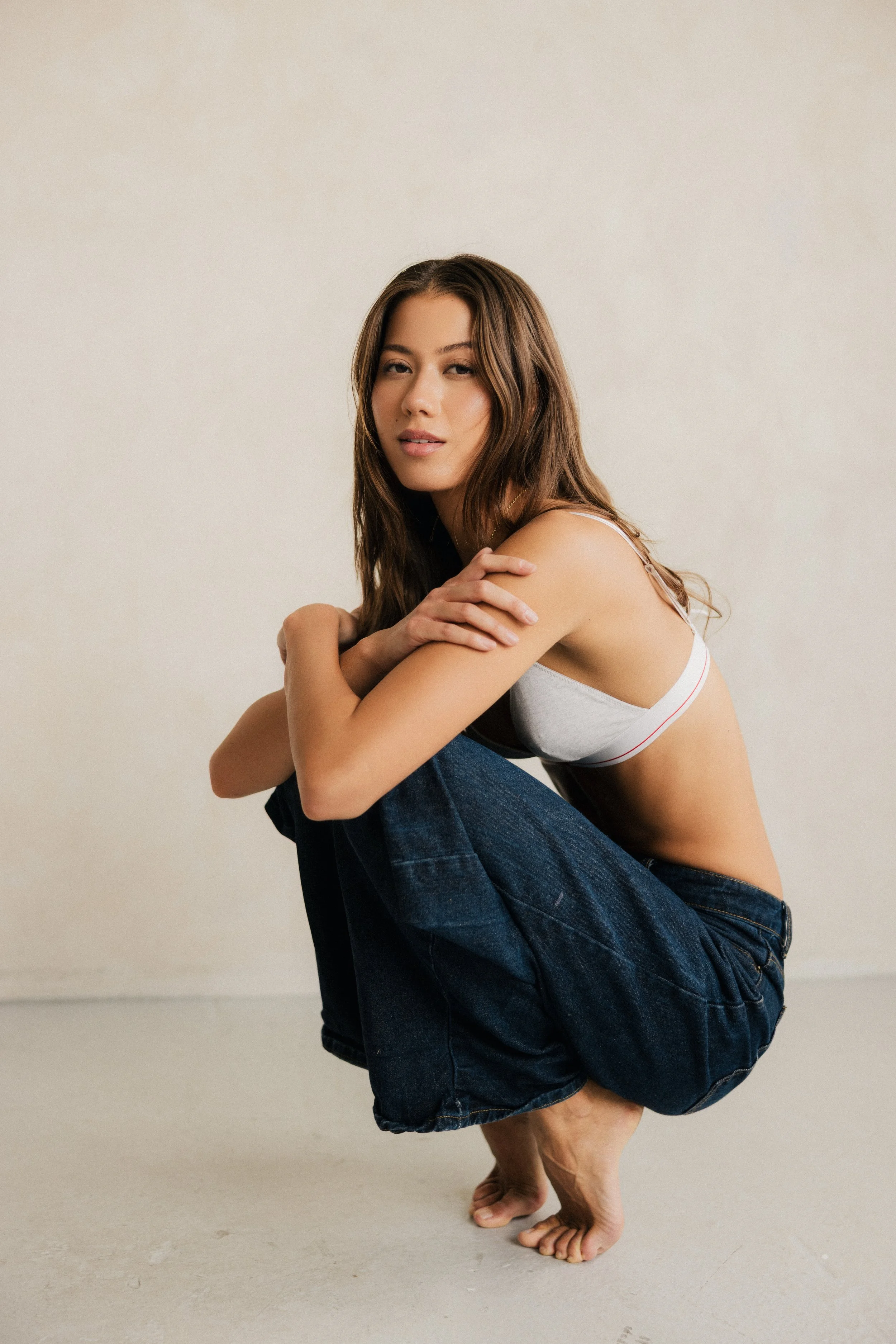 A young woman with long brown hair sitting on the shoulders of a man, with her arms crossed over her knees and wearing casual clothing, against a plain wall.