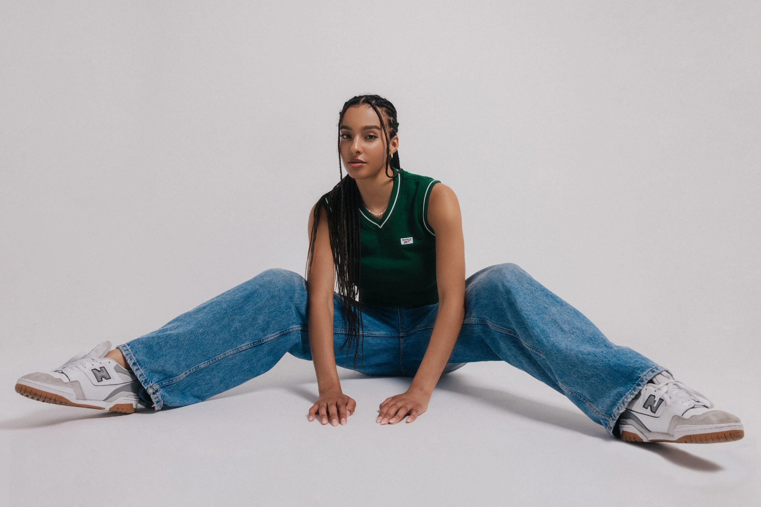 A young woman with long braided hair sitting on the floor with her legs spread apart and her hands on the ground. She wears a green sleeveless top, blue jeans, and white sneakers, posed against a plain white background.