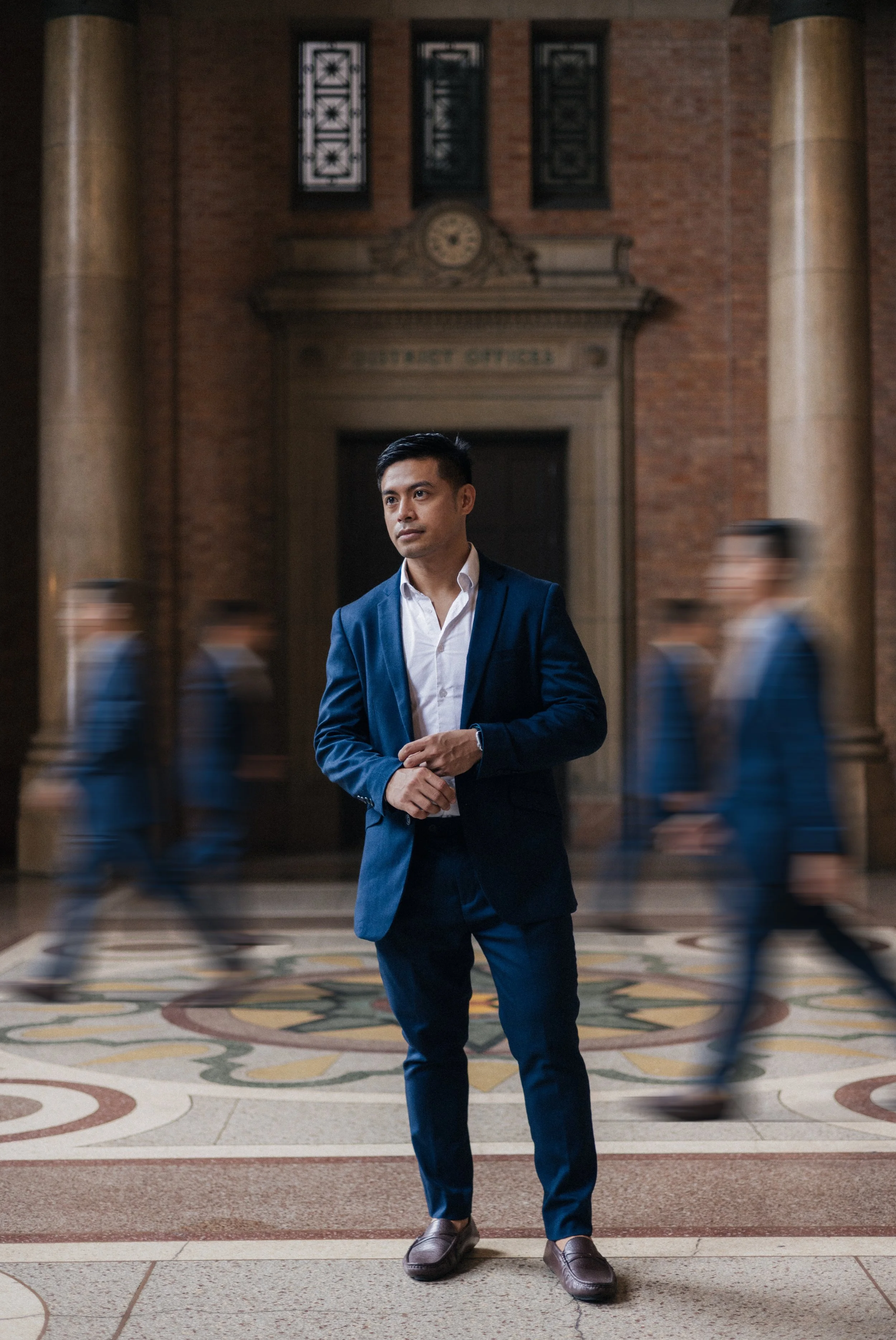 A man in a blue suit stands in a grand hallway with a decorative fireplace and stained glass windows, with blurred figures walking by.