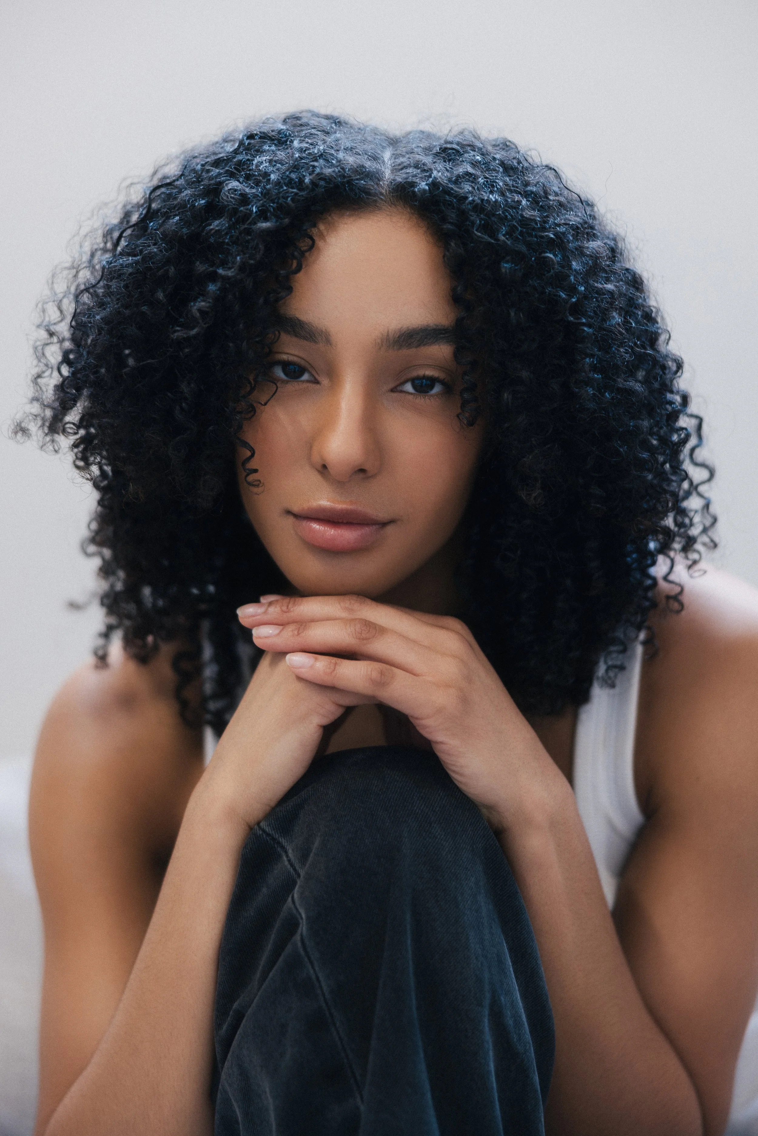 A woman with dark, curly hair and light brown skin sitting with her chin resting on her folded hands, looking into the camera