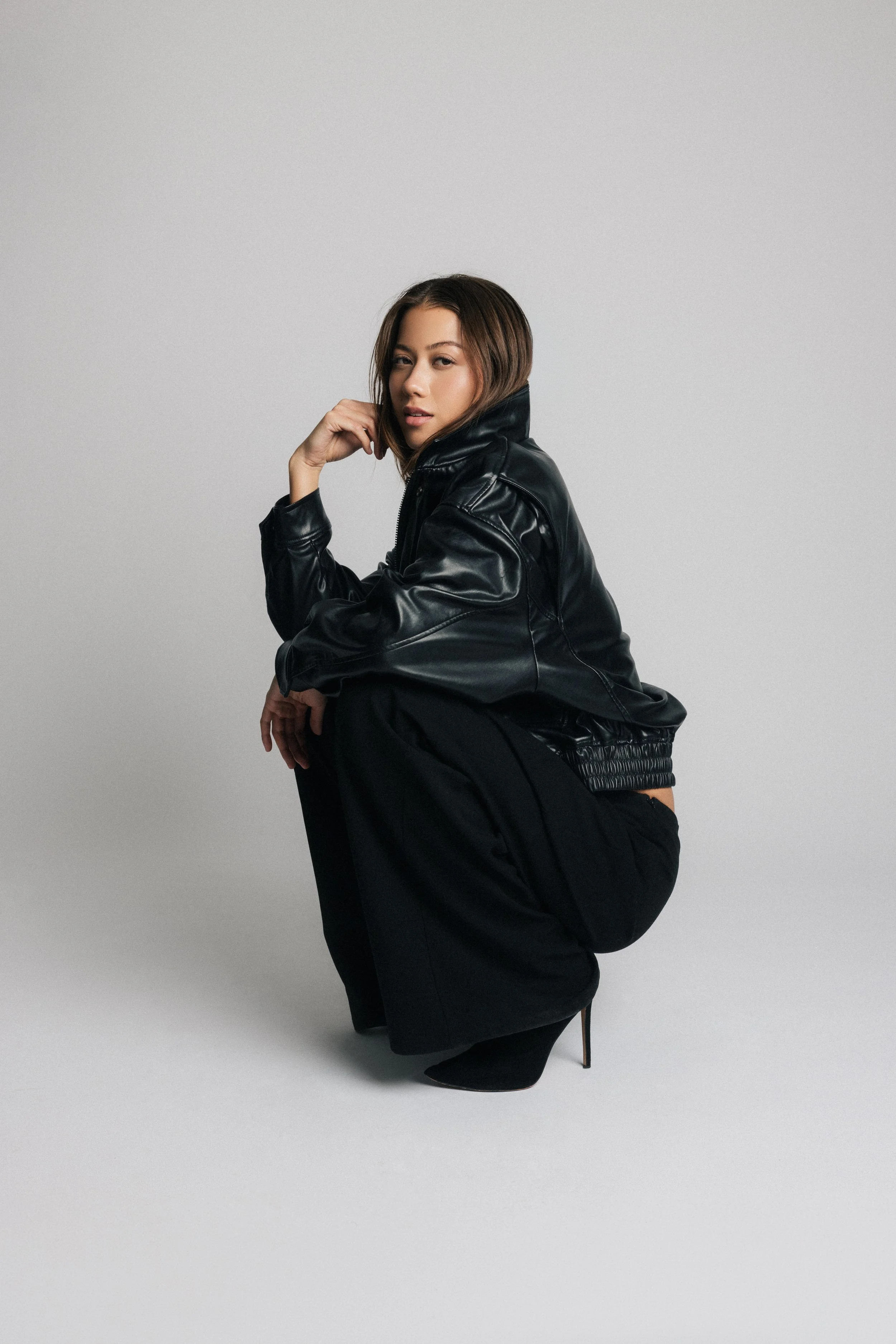 A woman wearing a black leather jacket and black high heeled shoes, squatting on the floor, with one hand near her face and the other resting on her knee, posing against a plain gray background.