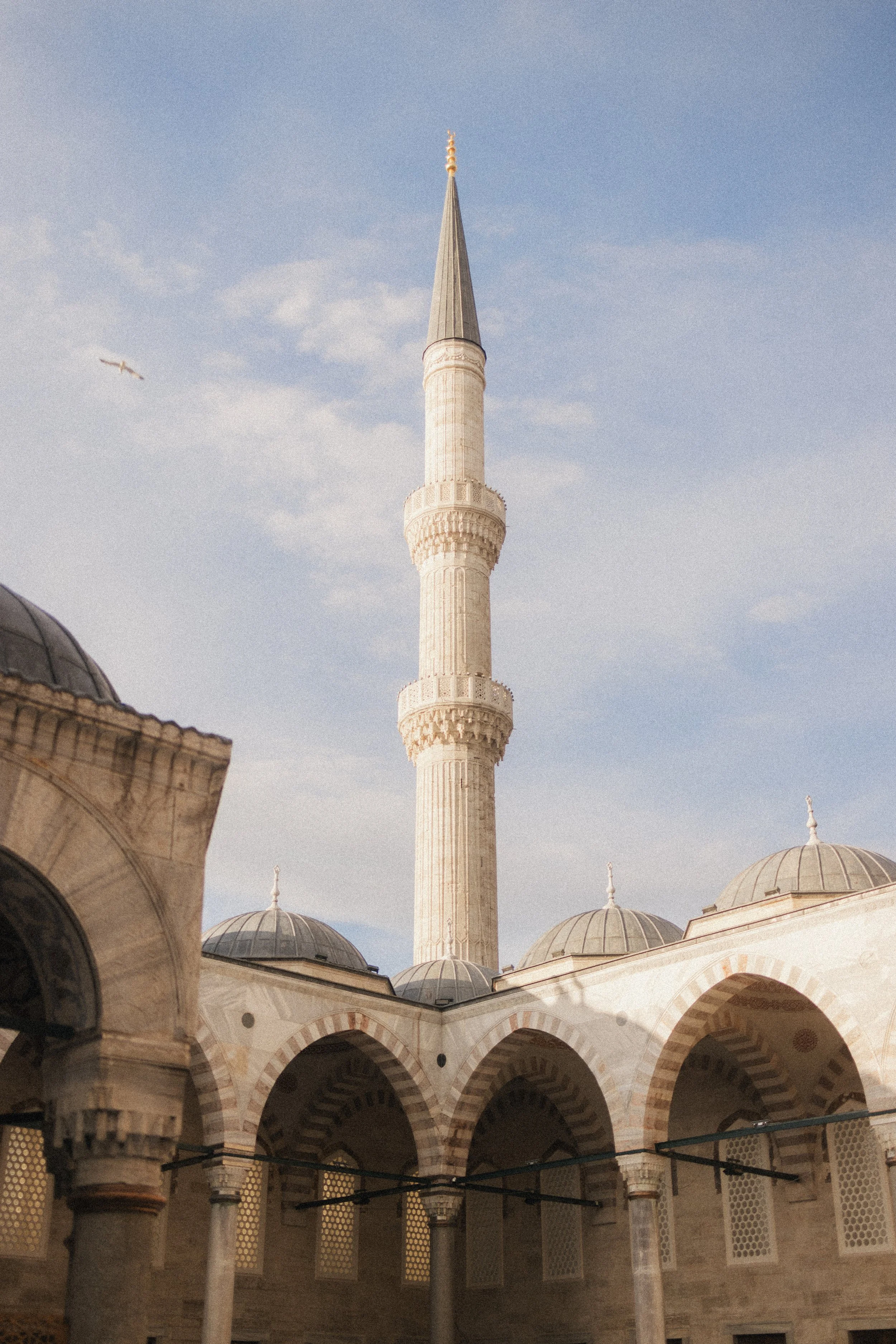View of a mosque's courtyard with arches and domes, a tall minaret, and a partly cloudy sky in the background.