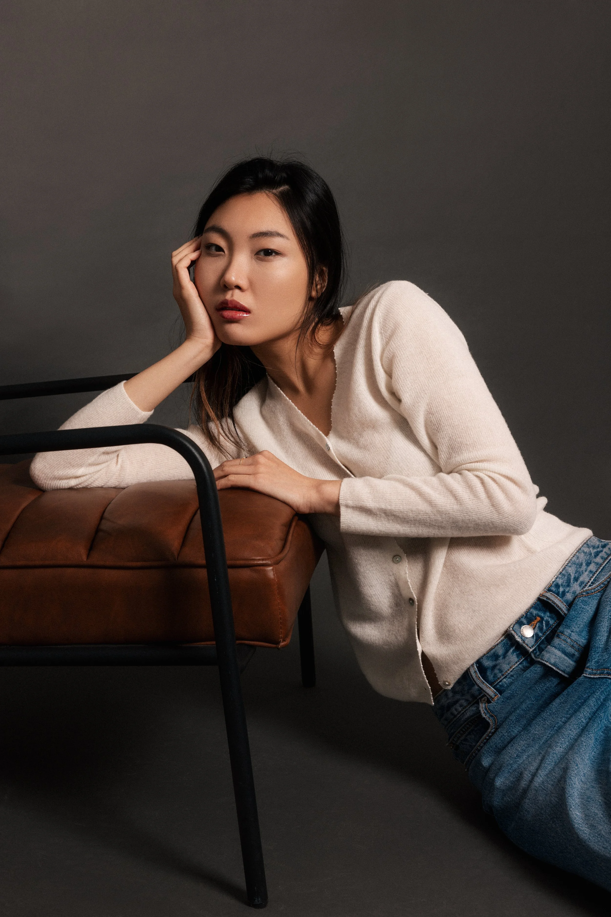 A young woman with black hair, wearing a cream cardigan and blue jeans, resting her head on her hand while leaning on a brown leather ottoman against a gray background.
