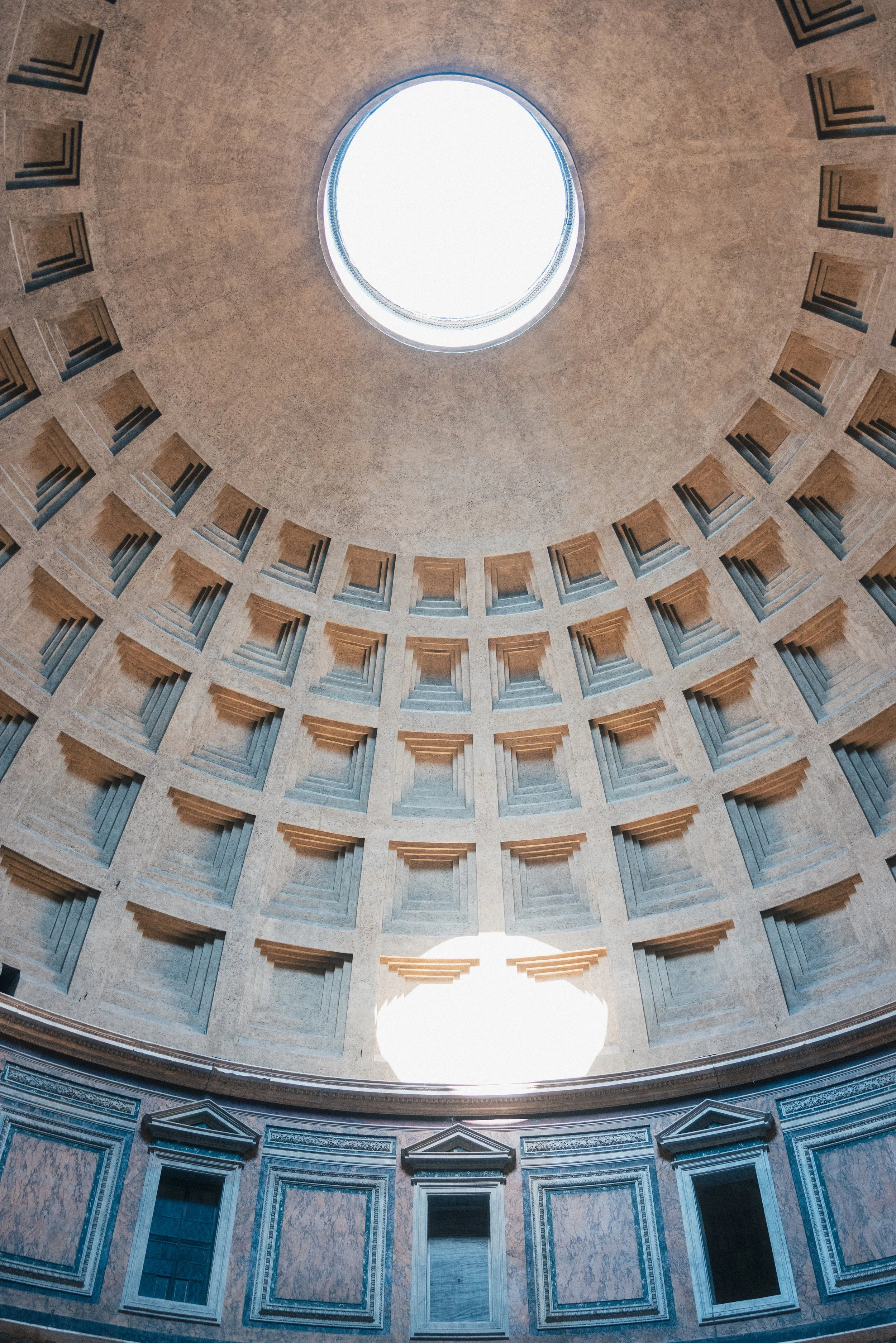 Interior view of the dome of the Pantheon showing the oculus at the top with sunlight streaming in, surrounded by geometric patterns and circles.
