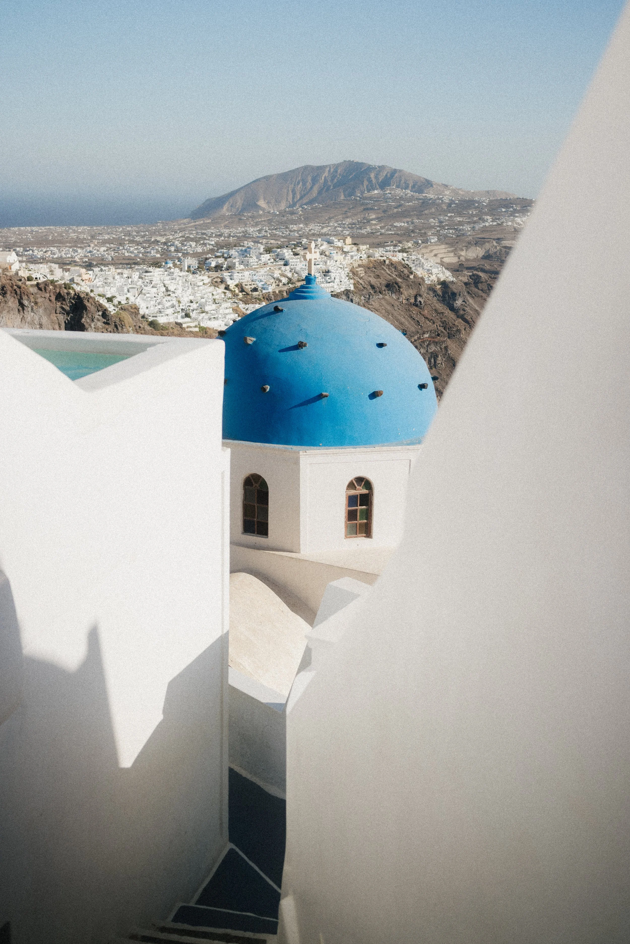 White building with blue domed roof, typical of Greek church architecture, against a backdrop of a town and mountain landscape.