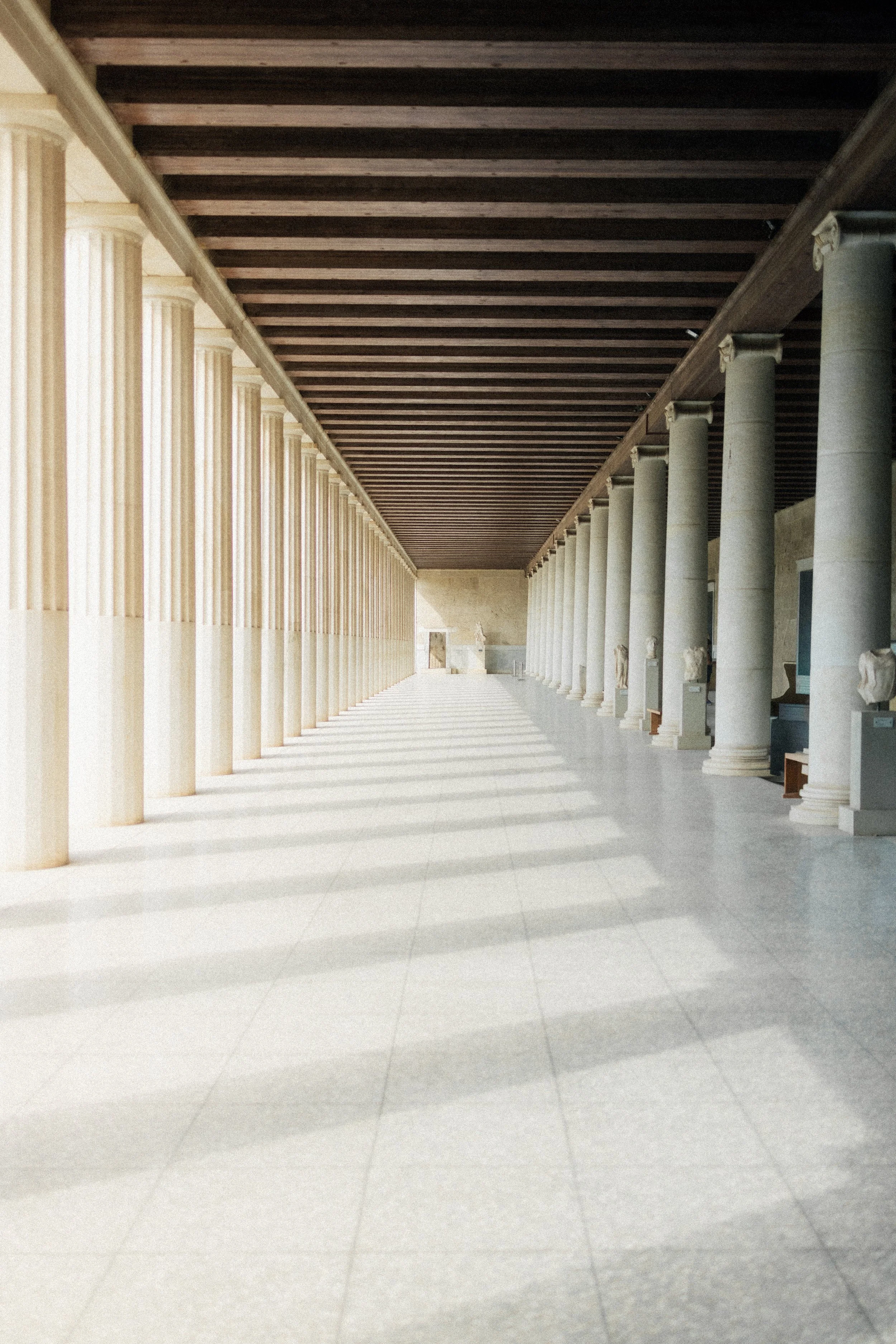 Long corridor with classical columns on both sides, a high dark wooden ceiling, and white tiled floor.