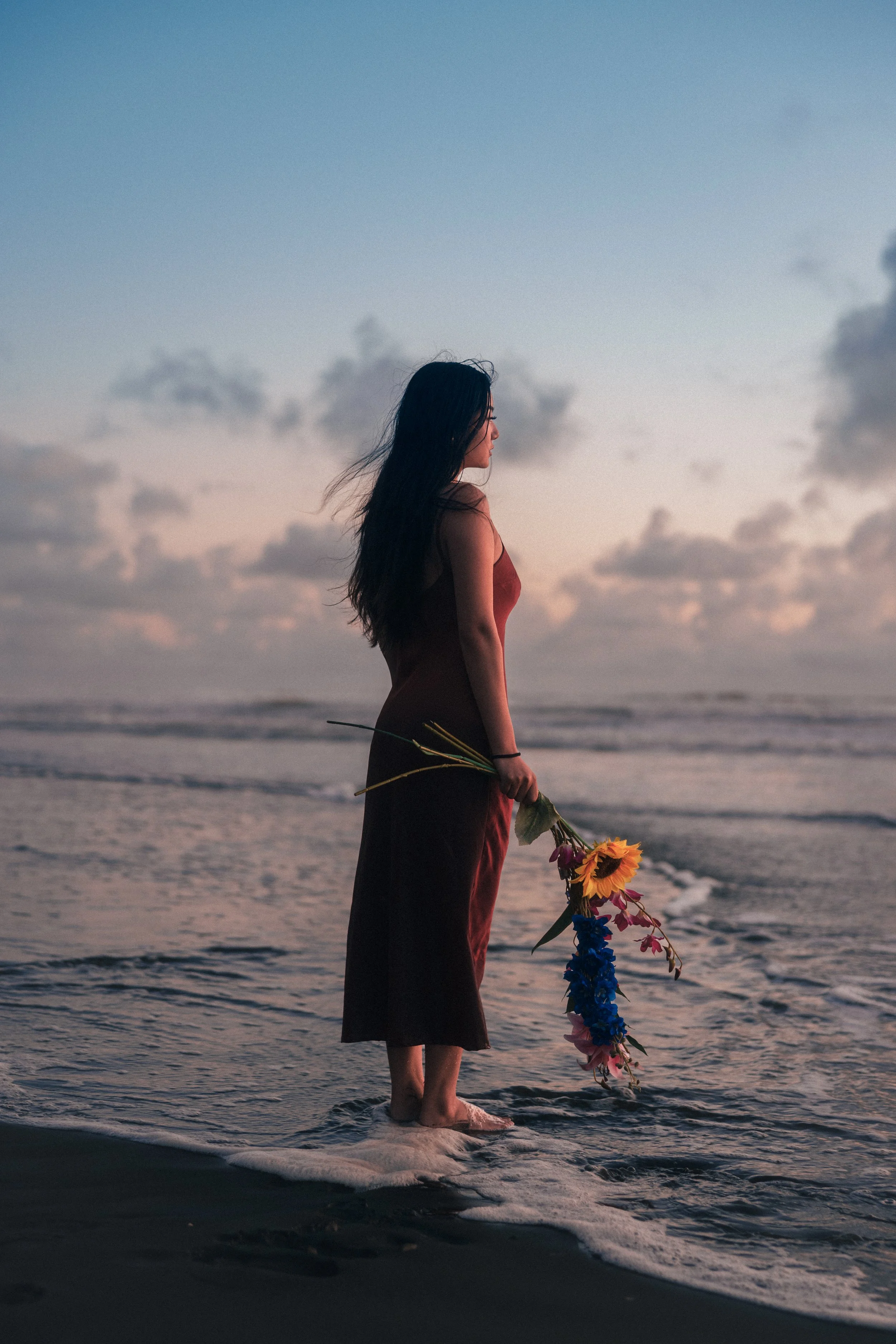 A woman with long dark hair standing at the edge of the ocean during sunset, holding a bouquet of colorful flowers, wearing a red dress.