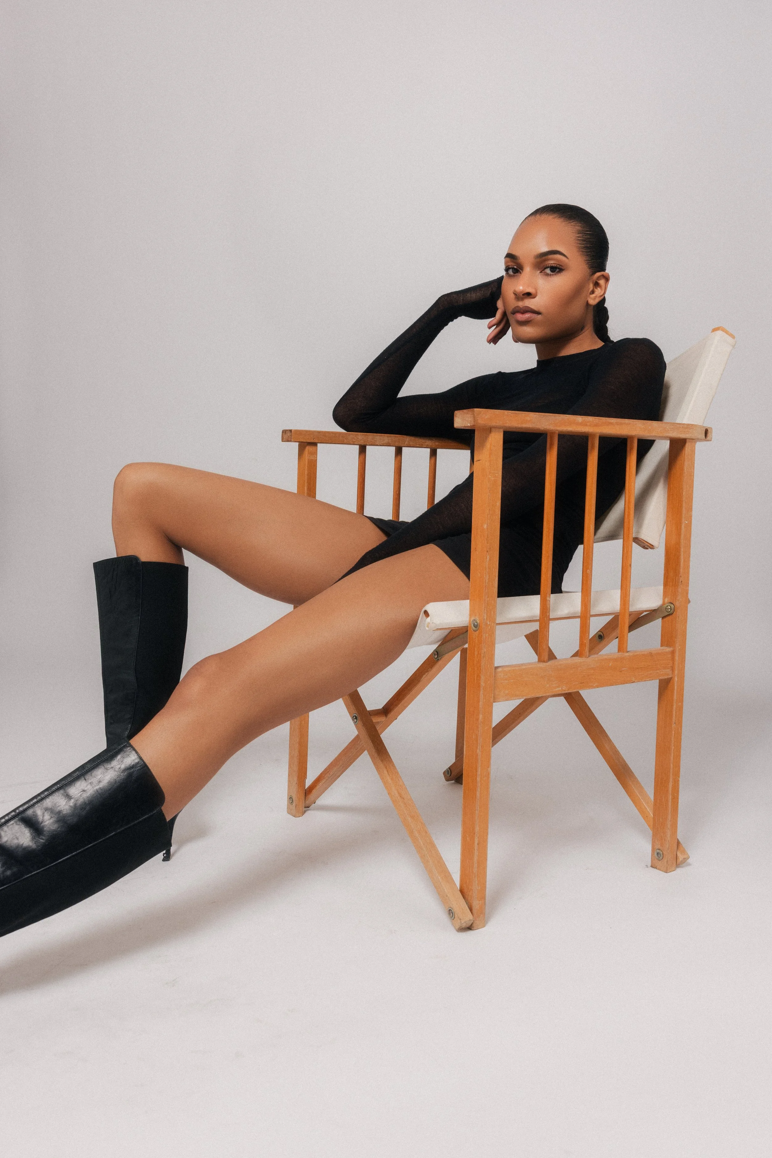 A woman with dark hair, wearing a black long sleeve top and black boots, sitting in a wooden chair with her legs extended, against a plain light gray background.