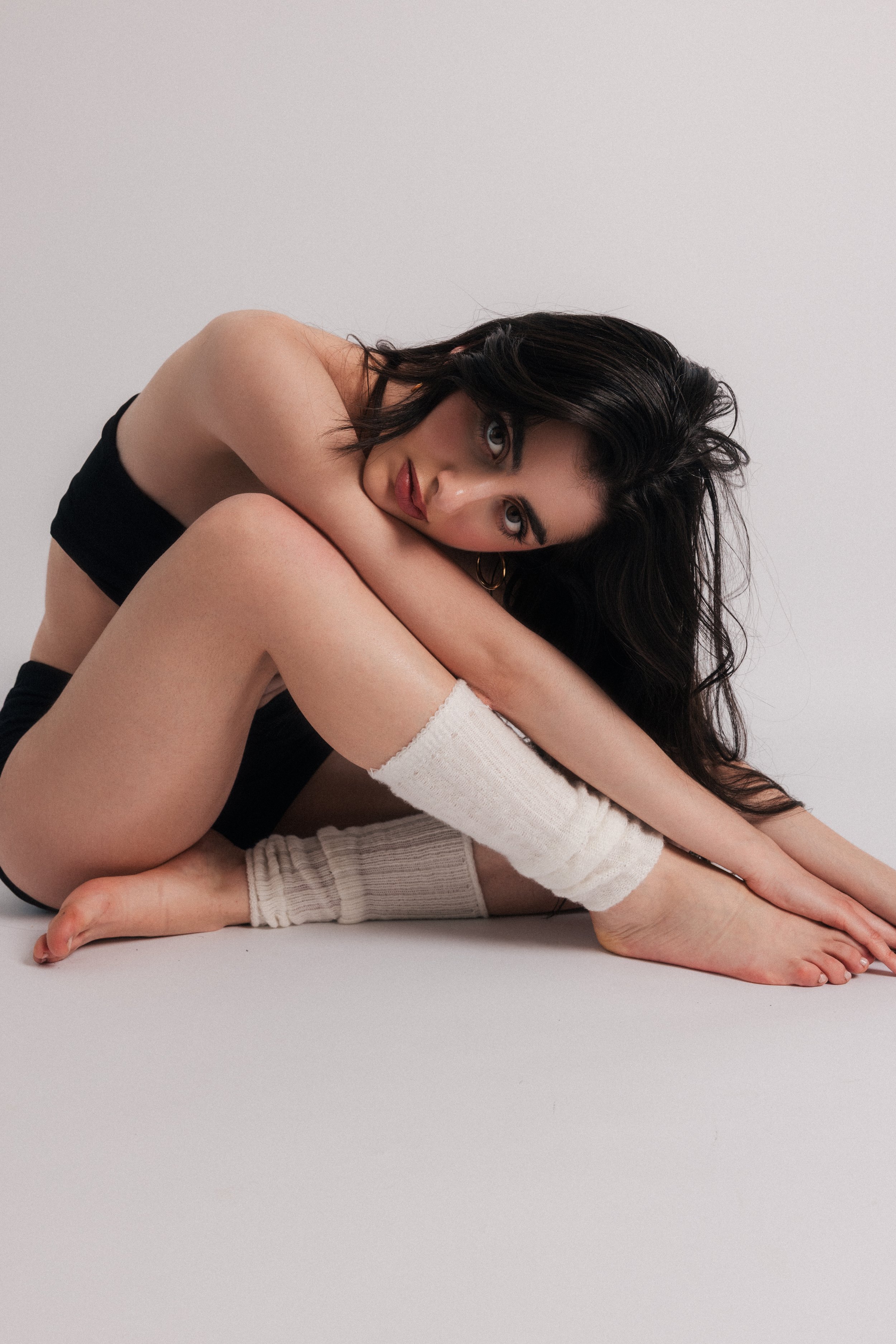 A woman with dark hair and tattoos sitting on the floor, wearing a black top and beige knee-high socks, against a plain white background.