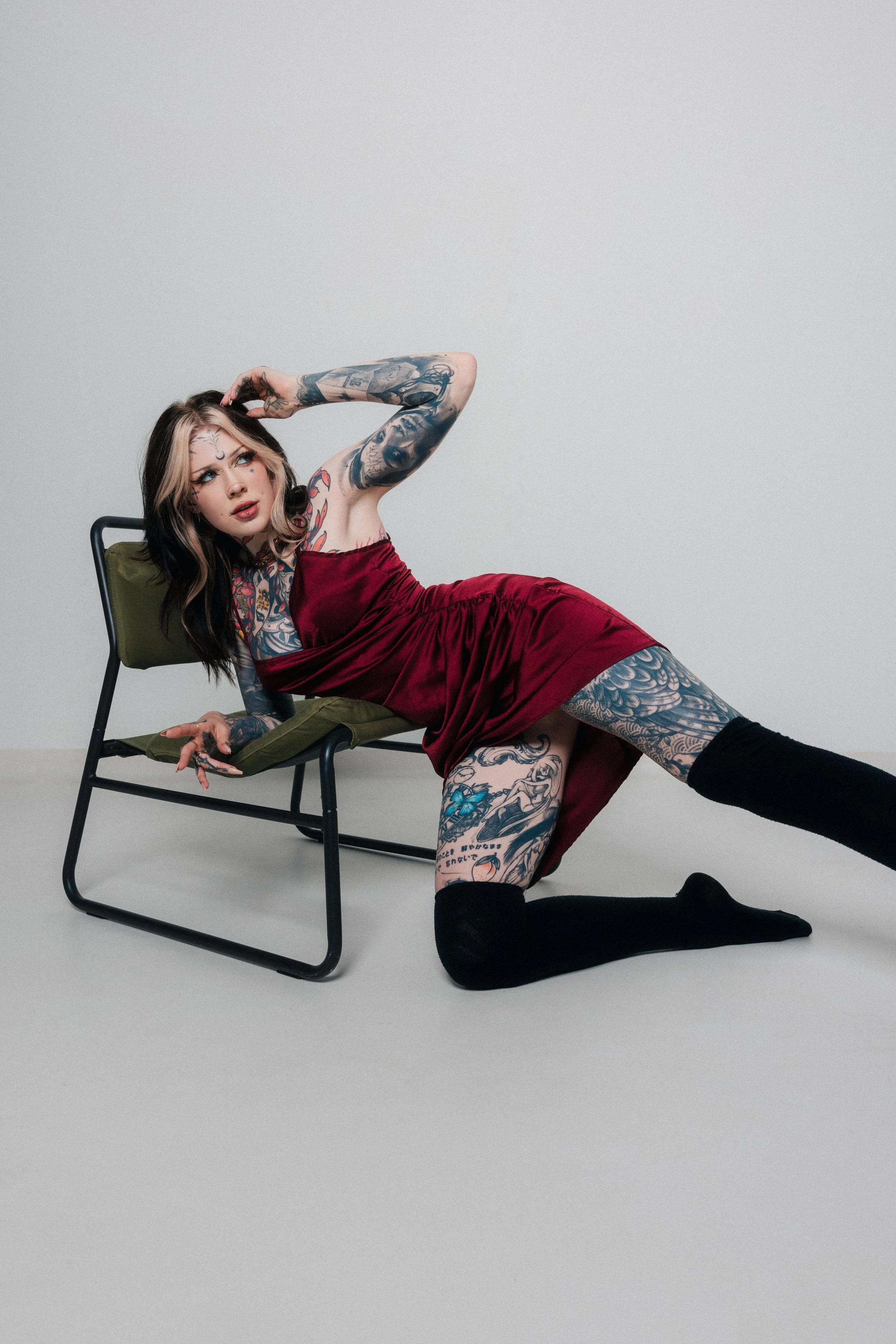 A woman with tattoos wearing a red dress, black thigh-high socks, and black slippers, posing on a gray floor with her head resting on a chair's armrest and her hand touching her head, against a plain gray background.