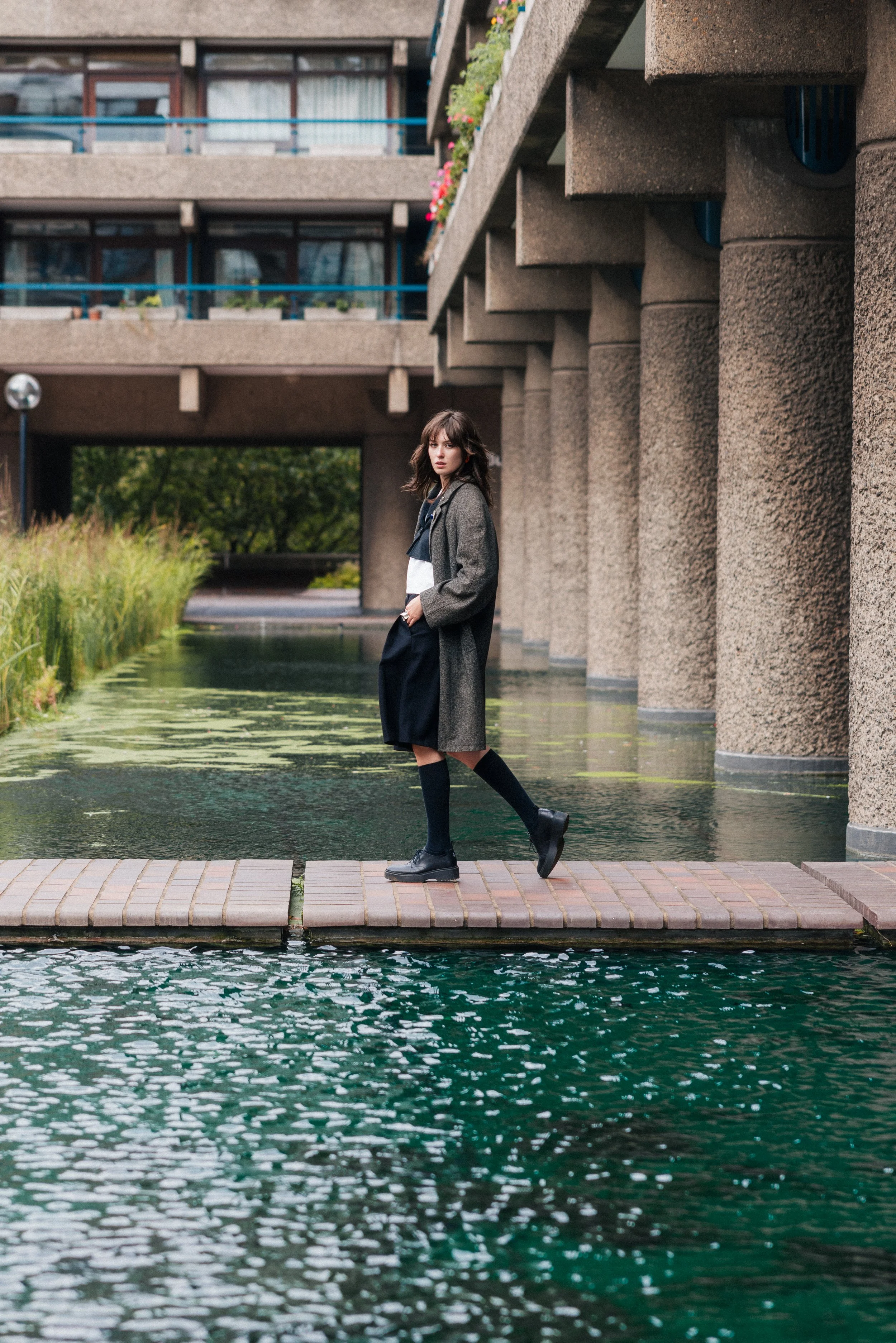A young woman with brown hair, wearing a gray coat, black top, black skirt, and black boots, standing on a small brick walkway over water. The background features a concrete building with balconies decorated with potted flowers and a canal lined with