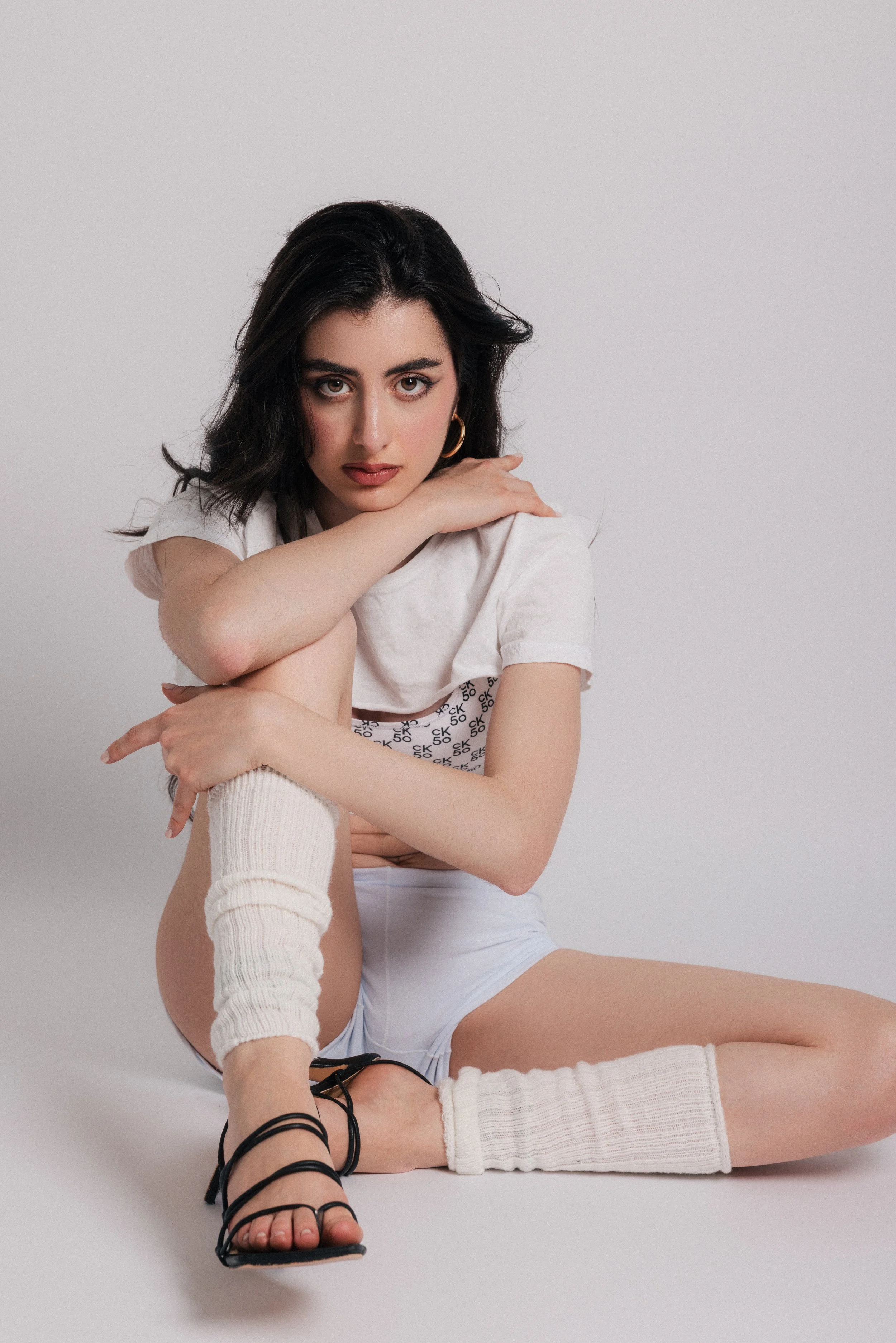A young woman with dark wavy hair, wearing a white T-shirt with 'CK 50' printed on it, white shorts, and thigh-high white knit socks, sitting on the floor with her arm resting on her knee, against a plain light grey background.