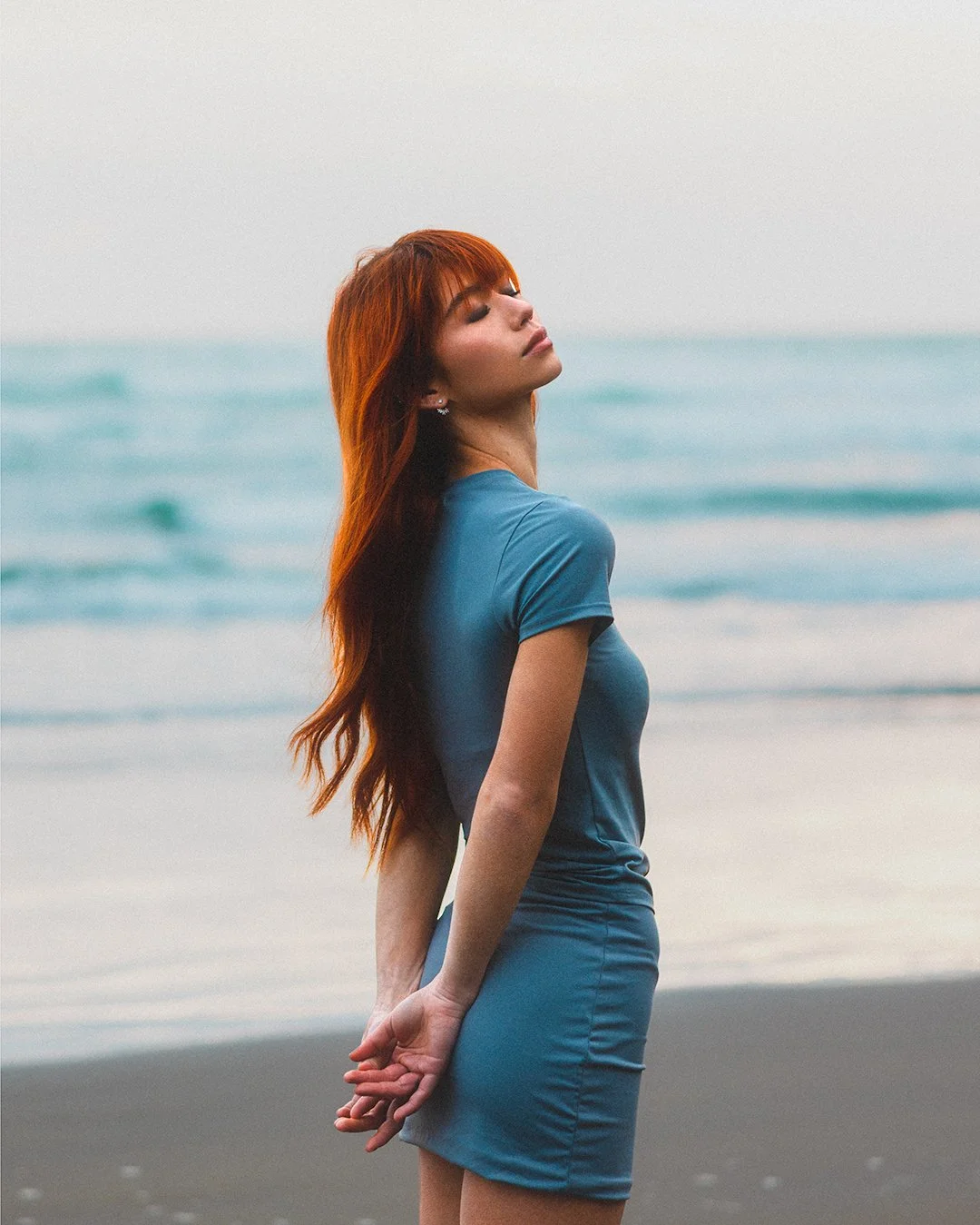 A woman with long red hair and earrings stands on the beach with her eyes closed, wearing a fitted blue dress, with the ocean in the background.